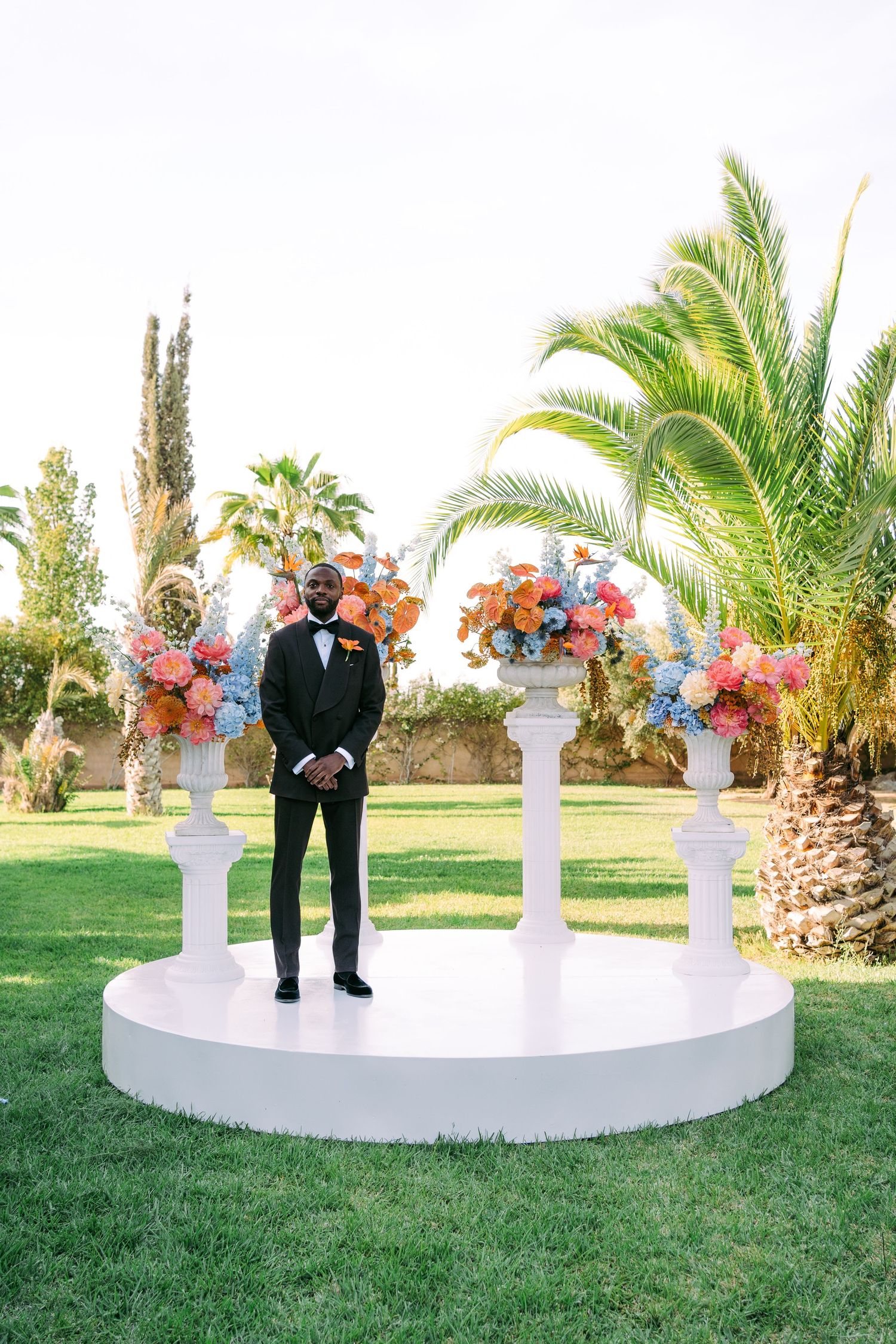 A wedding ceremony setup outdoors with white pedestals holding colorful floral arrangements against a palm tree backdrop.