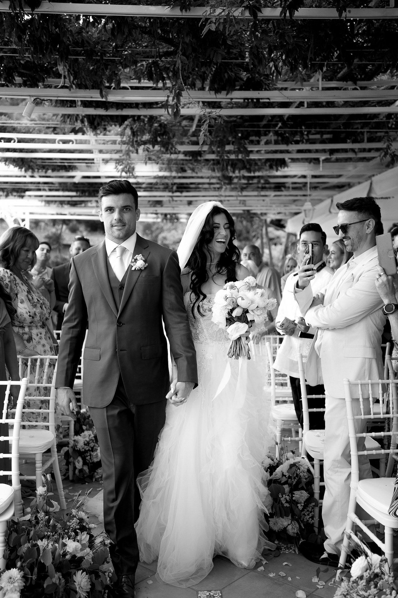 Wedding guests celebrate with confetti during an outdoor ceremony with white chairs and decorative arrangements.