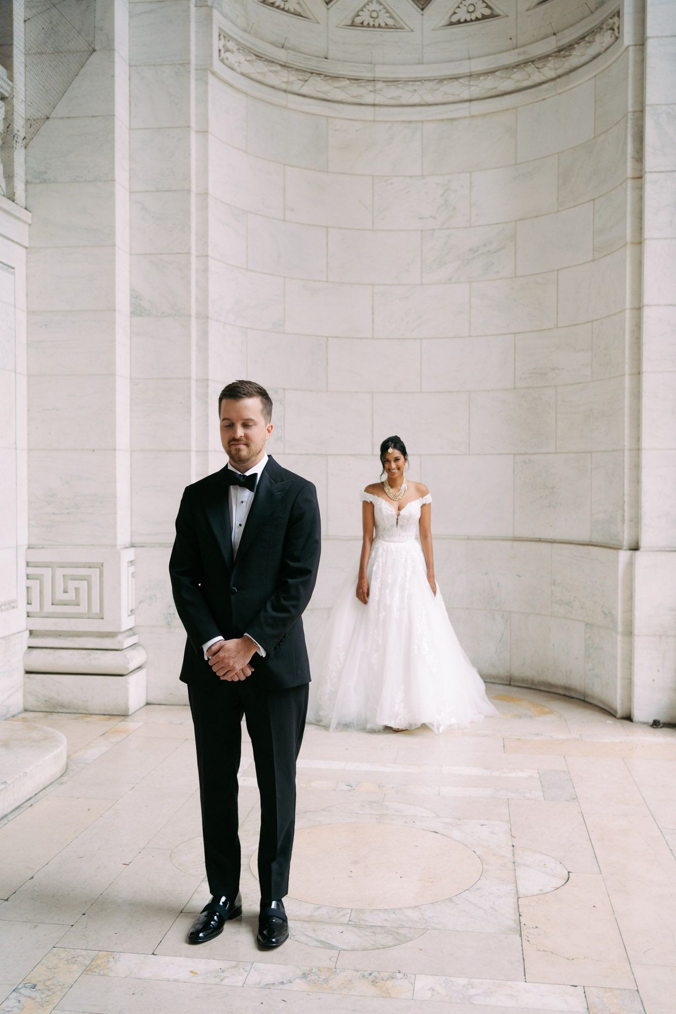 A couple in wedding attire poses in an elegant marble alcove with classical architectural details.