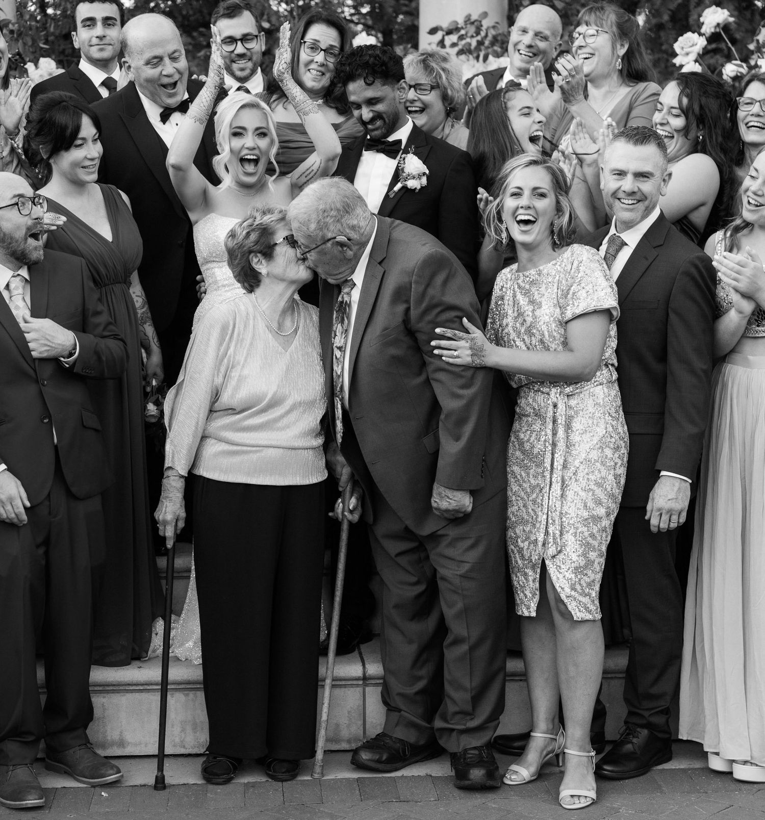 A large group of wedding guests laugh and smile together in a candid black and white photo.