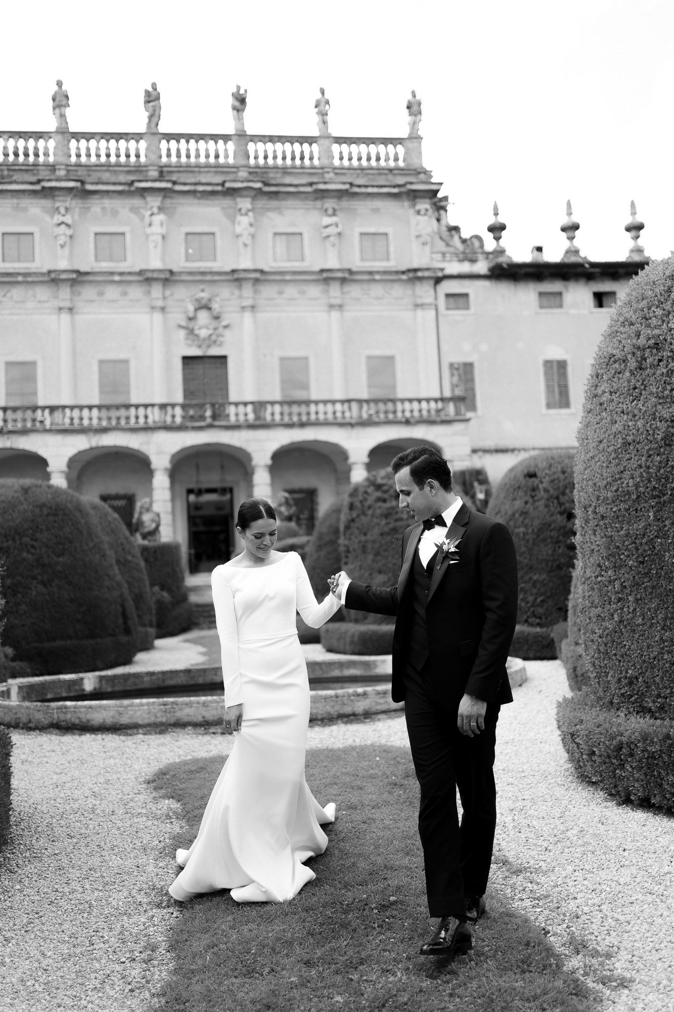Black and white photo of newlyweds posing in front of an ornate Italian villa with manicured hedges.