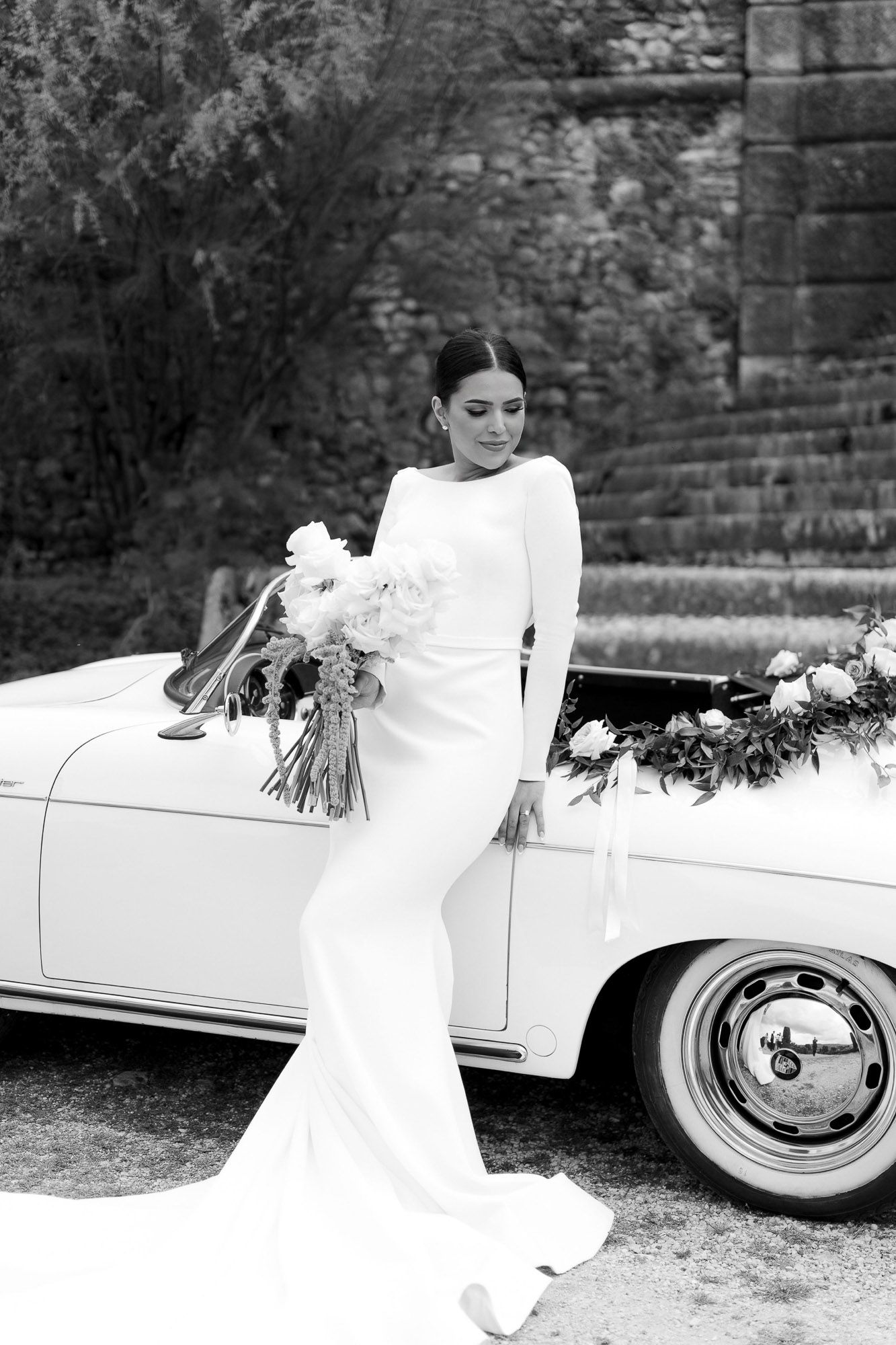 A vintage wedding scene with a classic white car decorated with flowers and a flowing white dress in black and white.