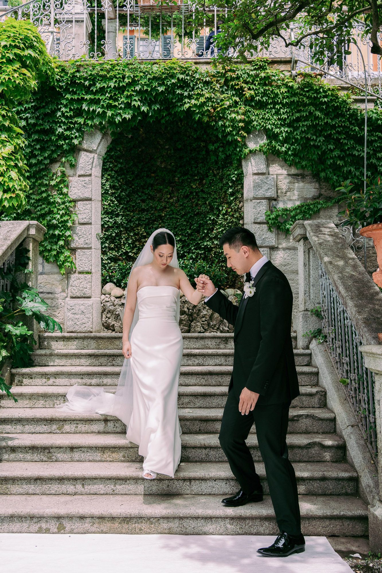 A couple in wedding attire shares a playful moment on stone steps surrounded by lush green ivy.