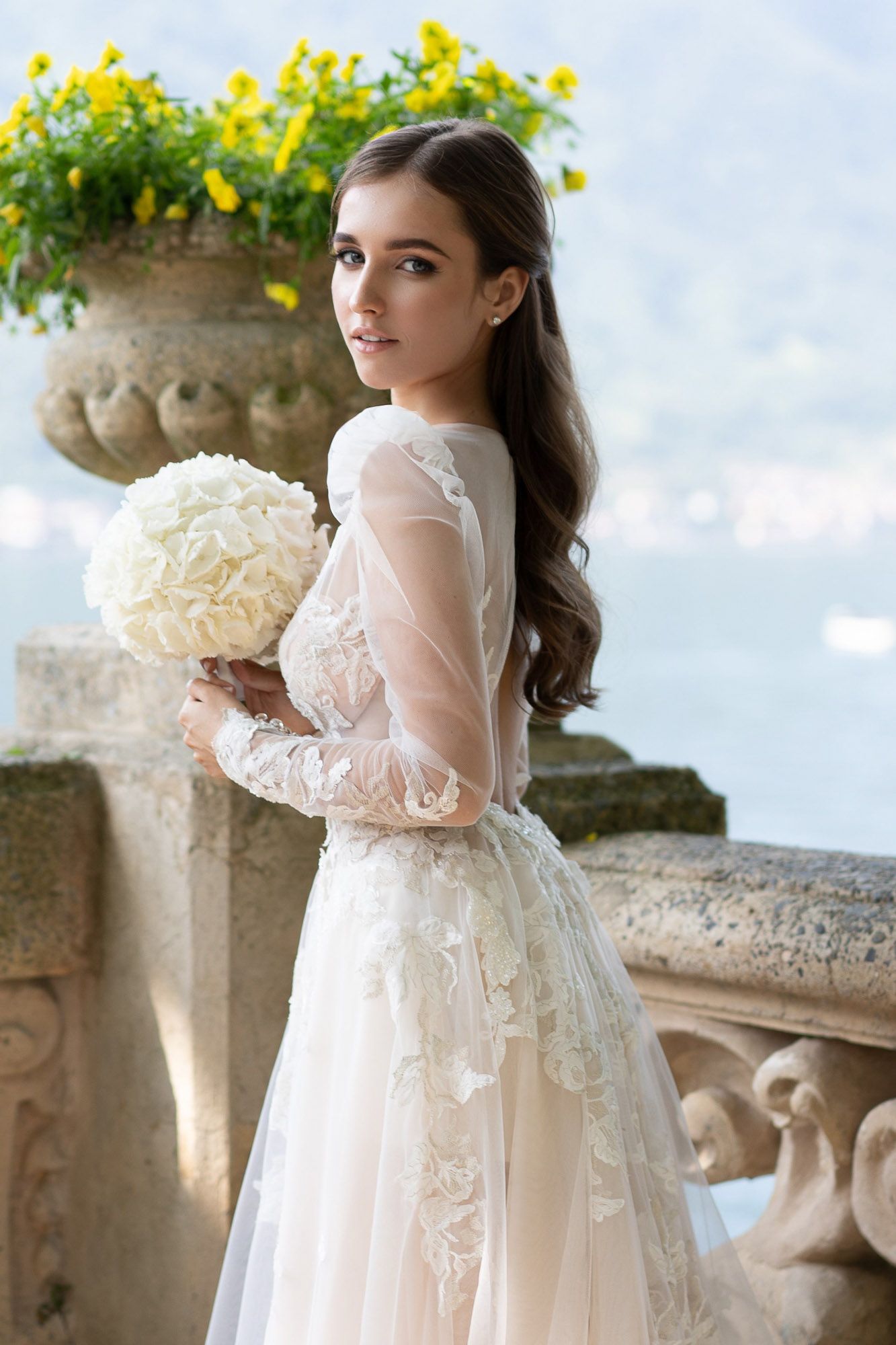 Elegant white lace dress with sheer sleeves photographed against a stone balcony with yellow flowers in the background.