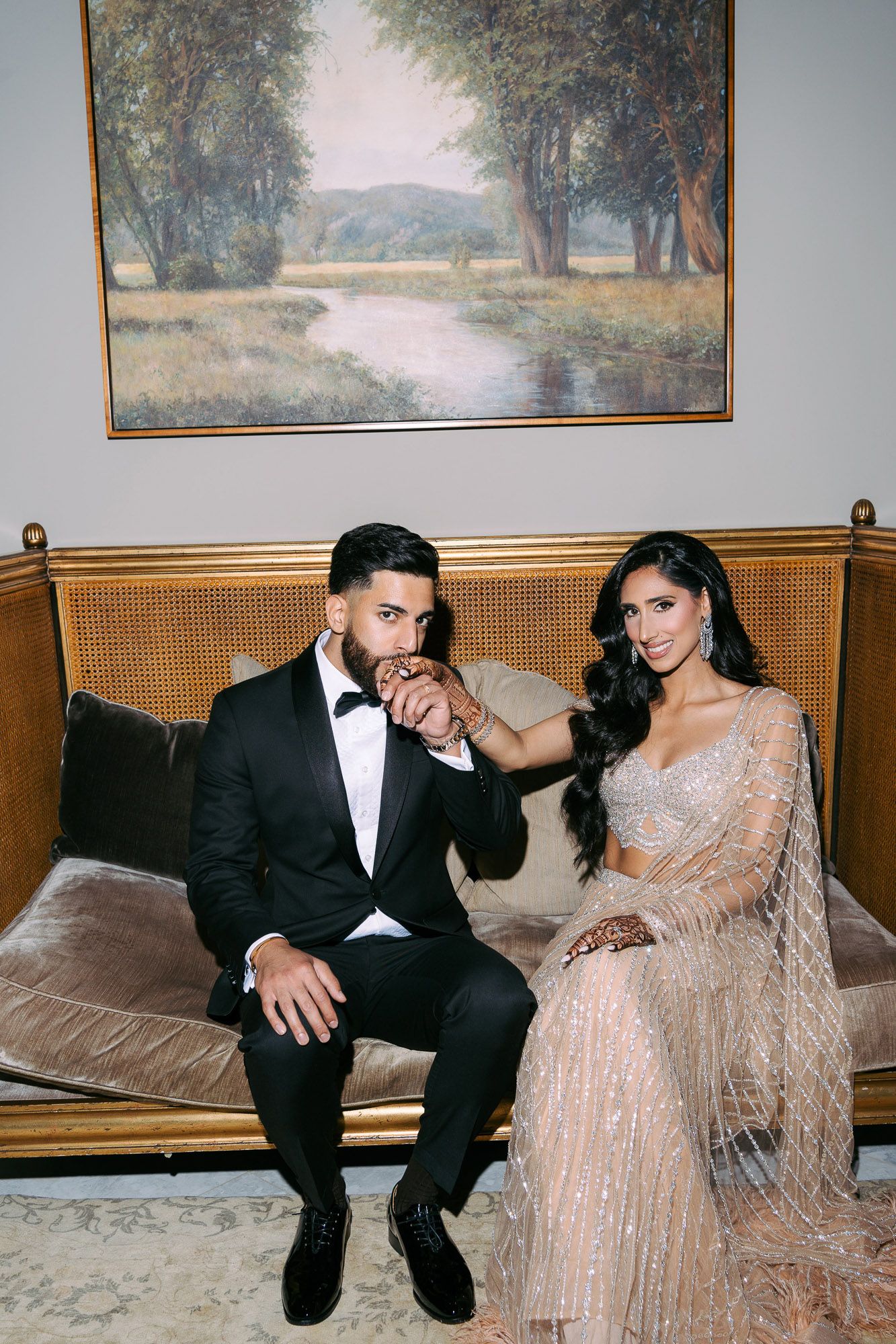 A Indian couple in evening wear poses intimately on a vintage bench with landscape paintings on the wall behind them.