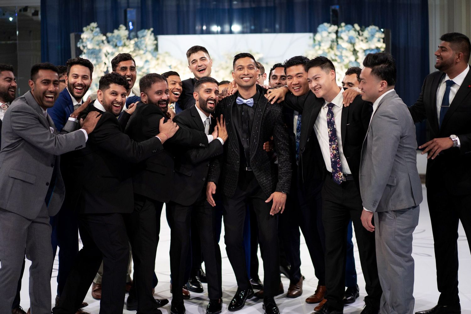 A group of well-dressed men in suits and tuxedos pose together at a formal event with decorative floral arrangements.