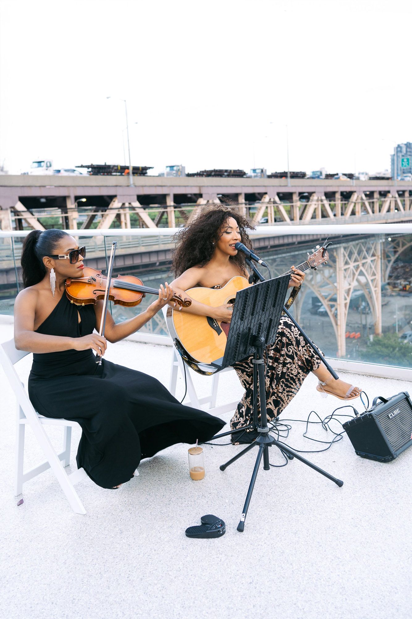 Two musicians perform with violin and guitar on a rooftop with a bridge visible in the background.