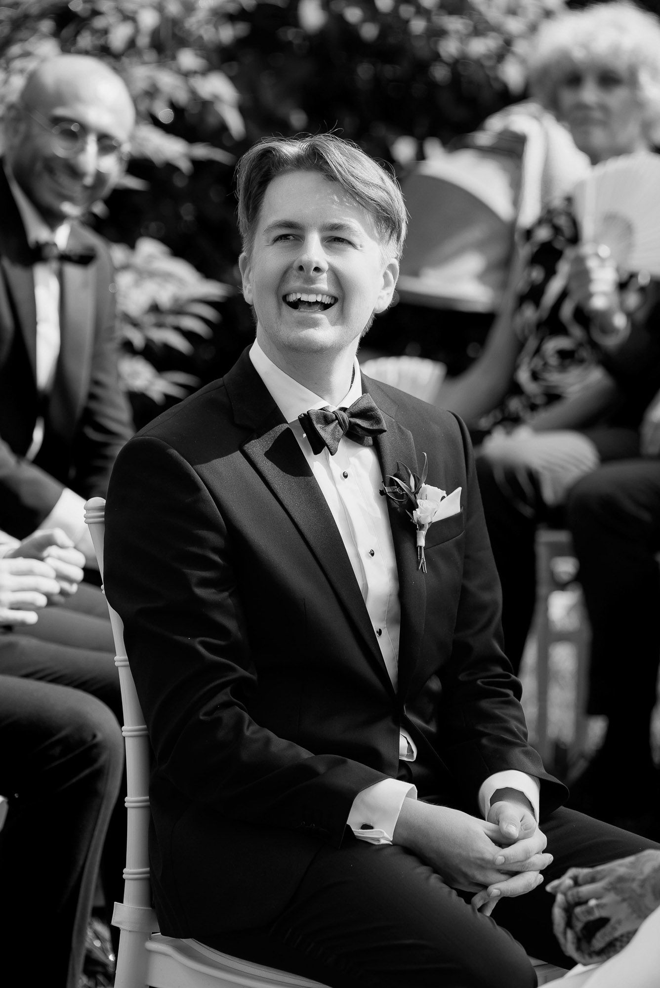 Young person in a black tuxedo sits and smiles during a formal event.