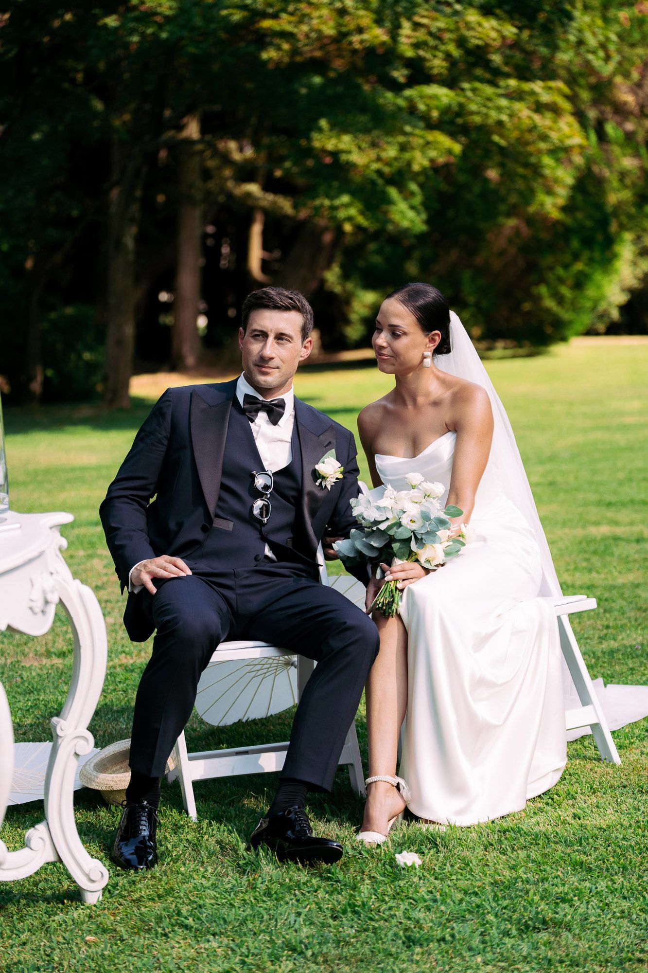 A couple in wedding attire sits together on white garden chairs on a lush green lawn.
