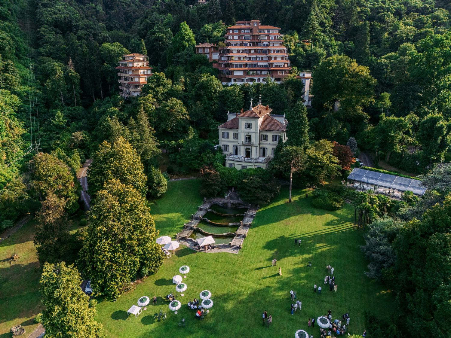 Aerial view of a historic mansion nestled in lush green gardens with parked cars and winding pathways.
