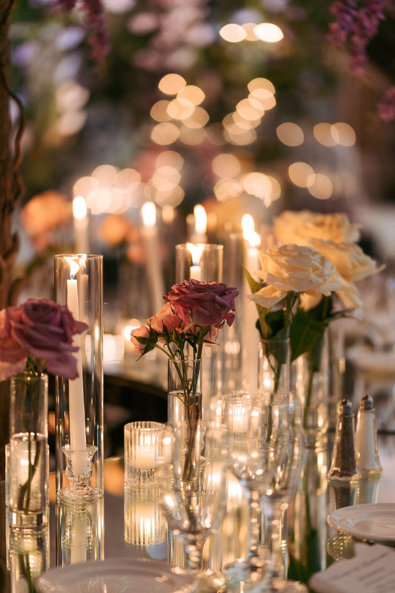 Elegant wedding reception table setting with glowing candles and purple roses in glass vases creating romantic ambiance.
