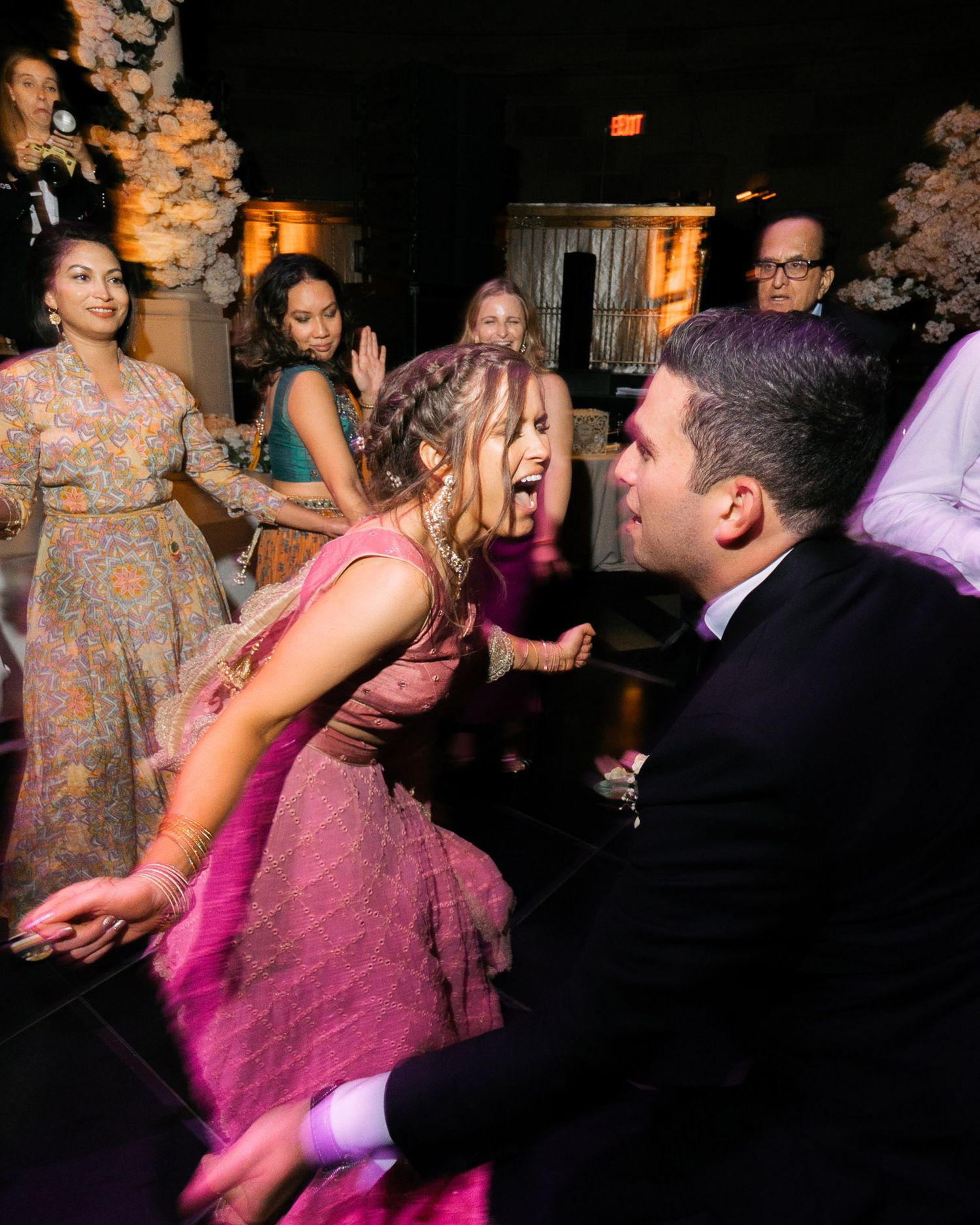 Wedding guests dance and celebrate on a dimly lit dance floor with warm lighting and festive atmosphere.