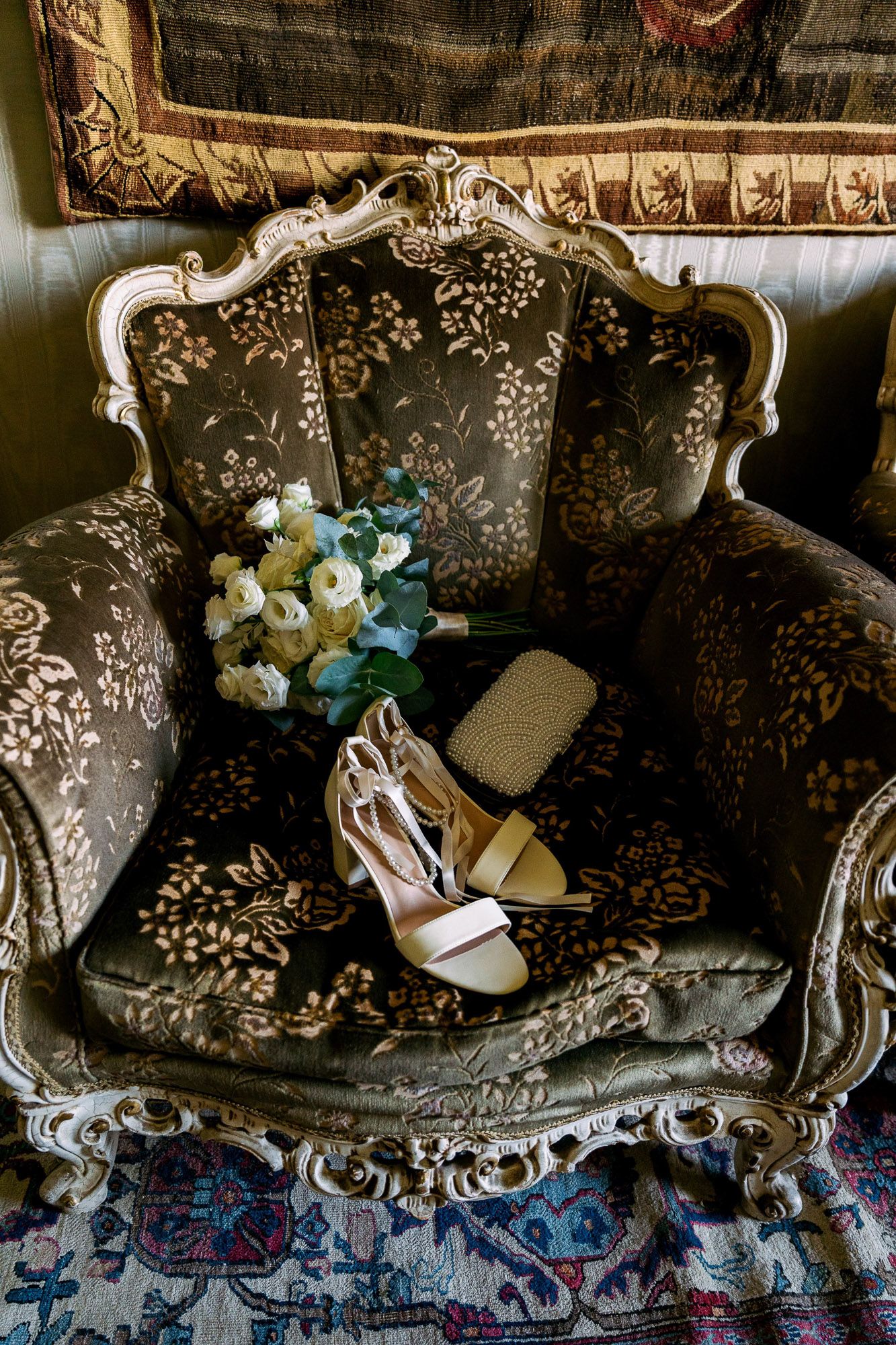 Wedding accessories including white heels and a floral bouquet arranged on an ornate vintage sofa with damask upholstery.