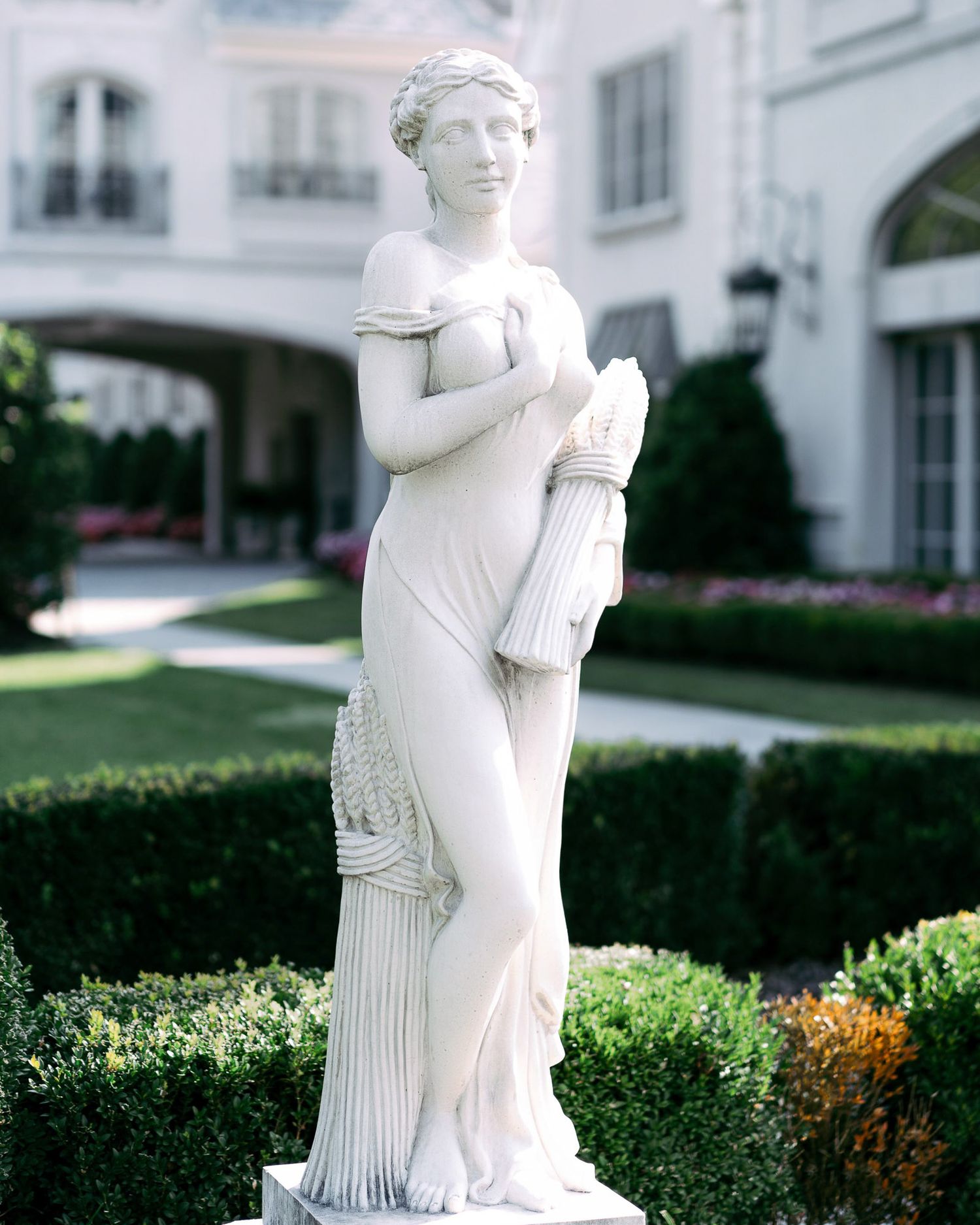 Classical white marble statues stand elegantly among manicured hedges in a formal garden setting.