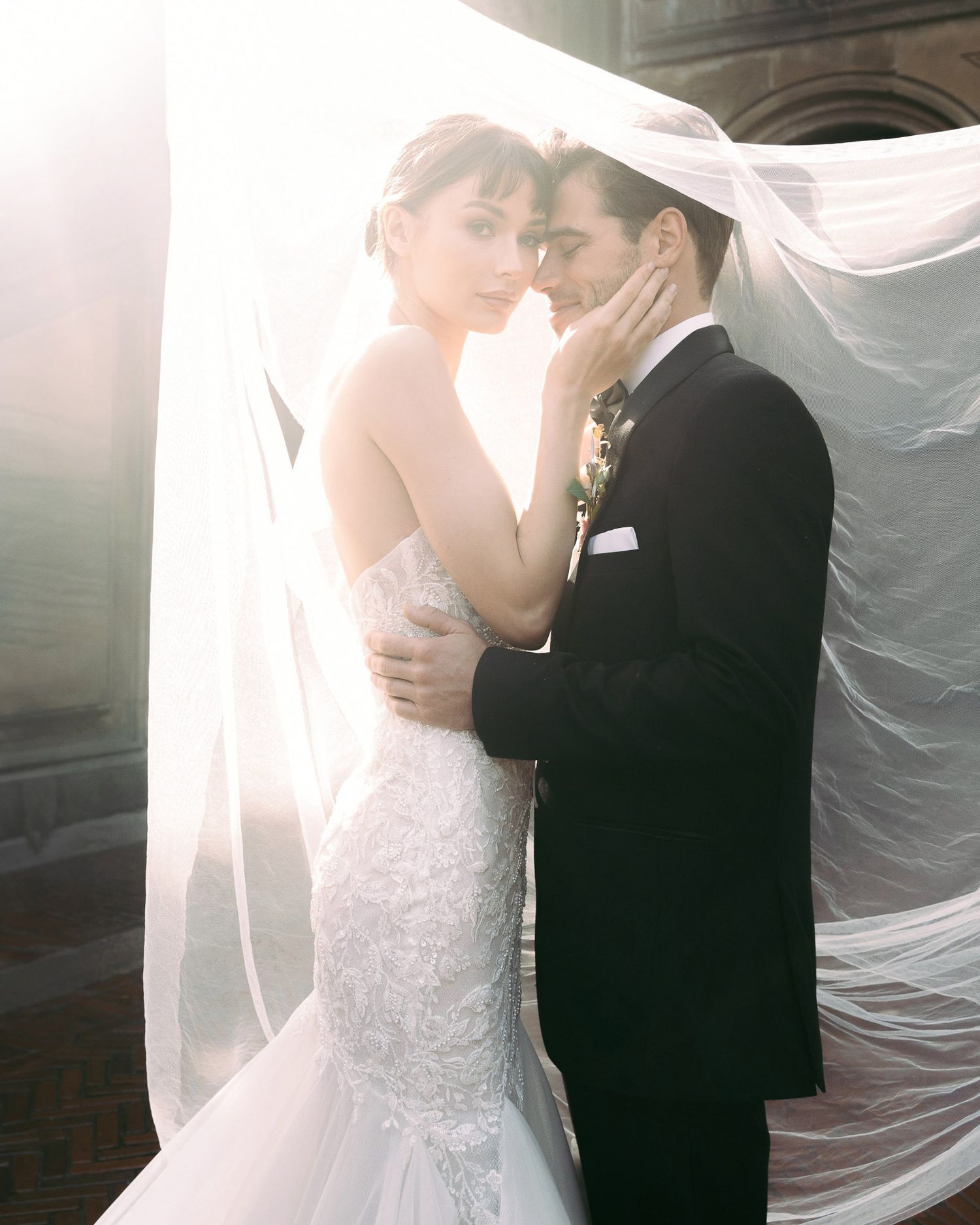A romantic wedding couple embraces in soft sunlight with a flowing veil creating an ethereal atmosphere.