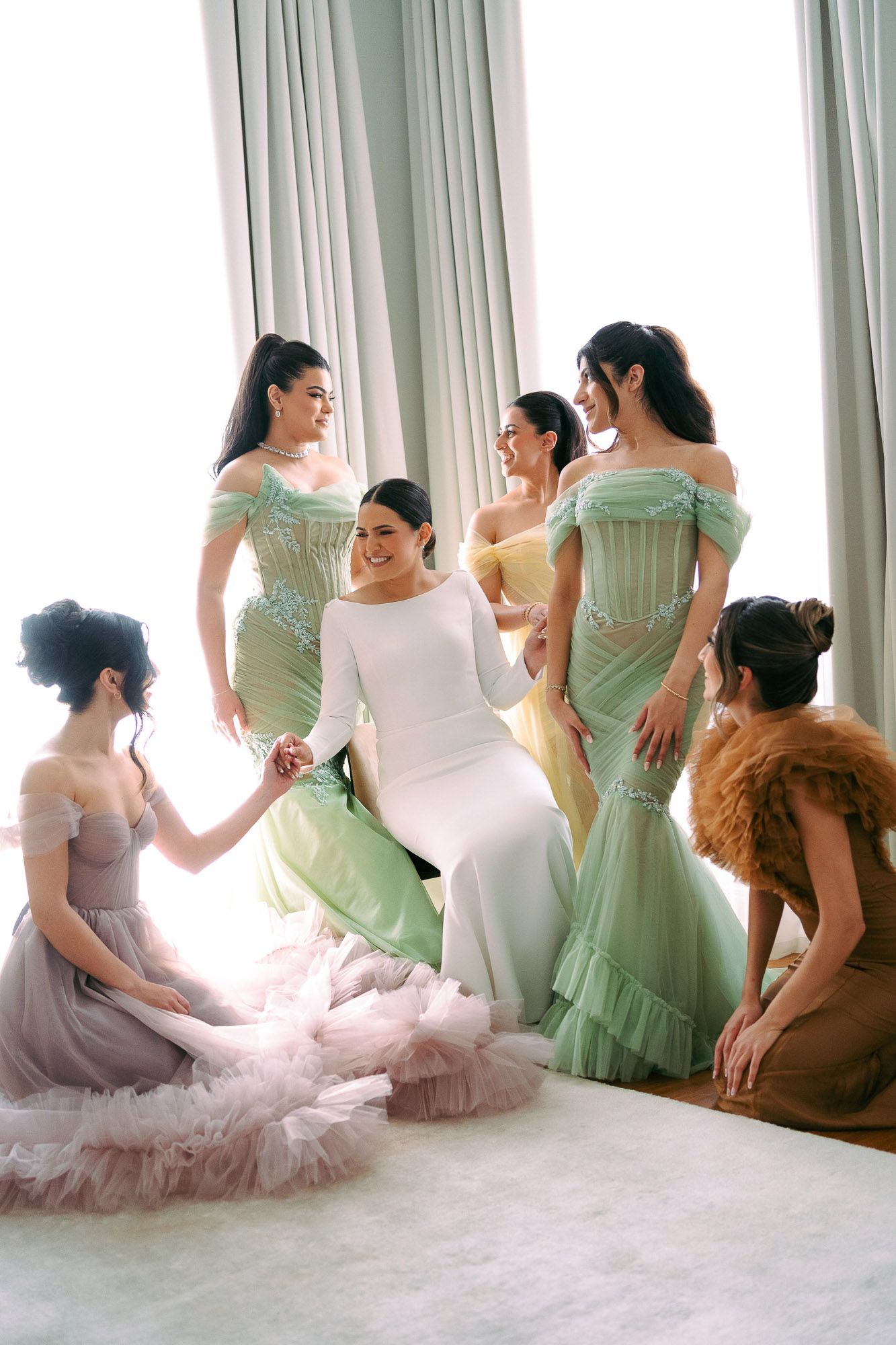 A bridal party in pastel mint green and lavender gowns gather together in an elegant room with flowing curtains.