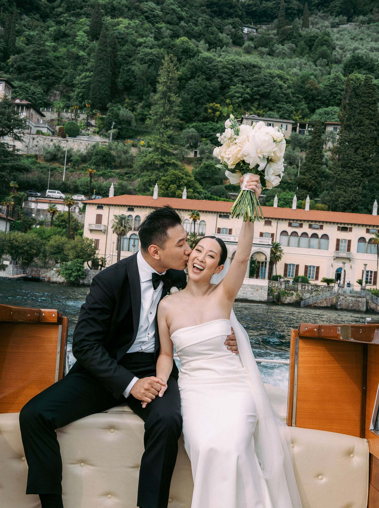 A romantic wedding couple shares intimate moments on a boat with Lake Como's picturesque Italian shoreline in the background.