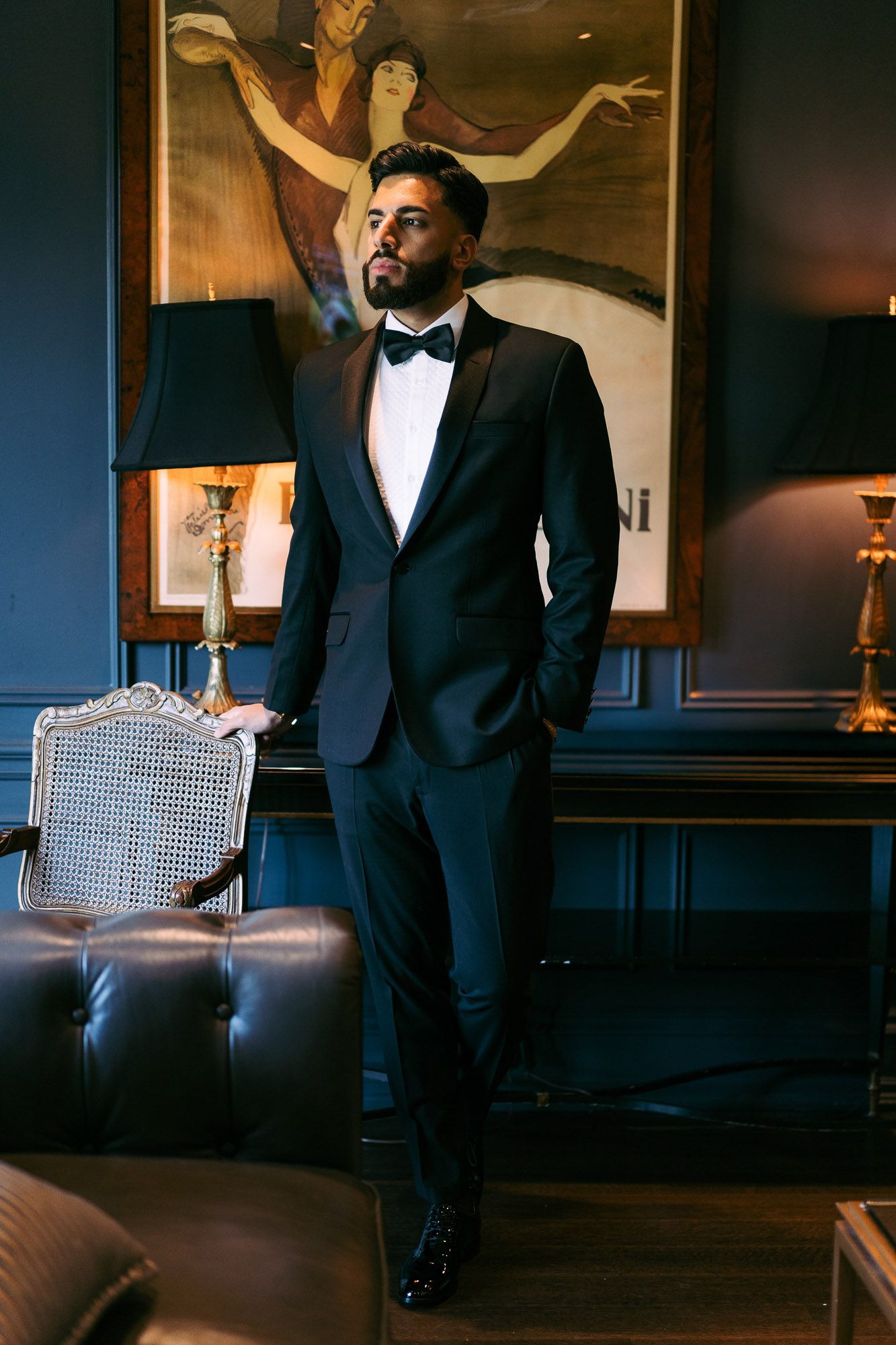 A person in a black tuxedo and bow tie poses dramatically in a dimly lit room with leather seating.