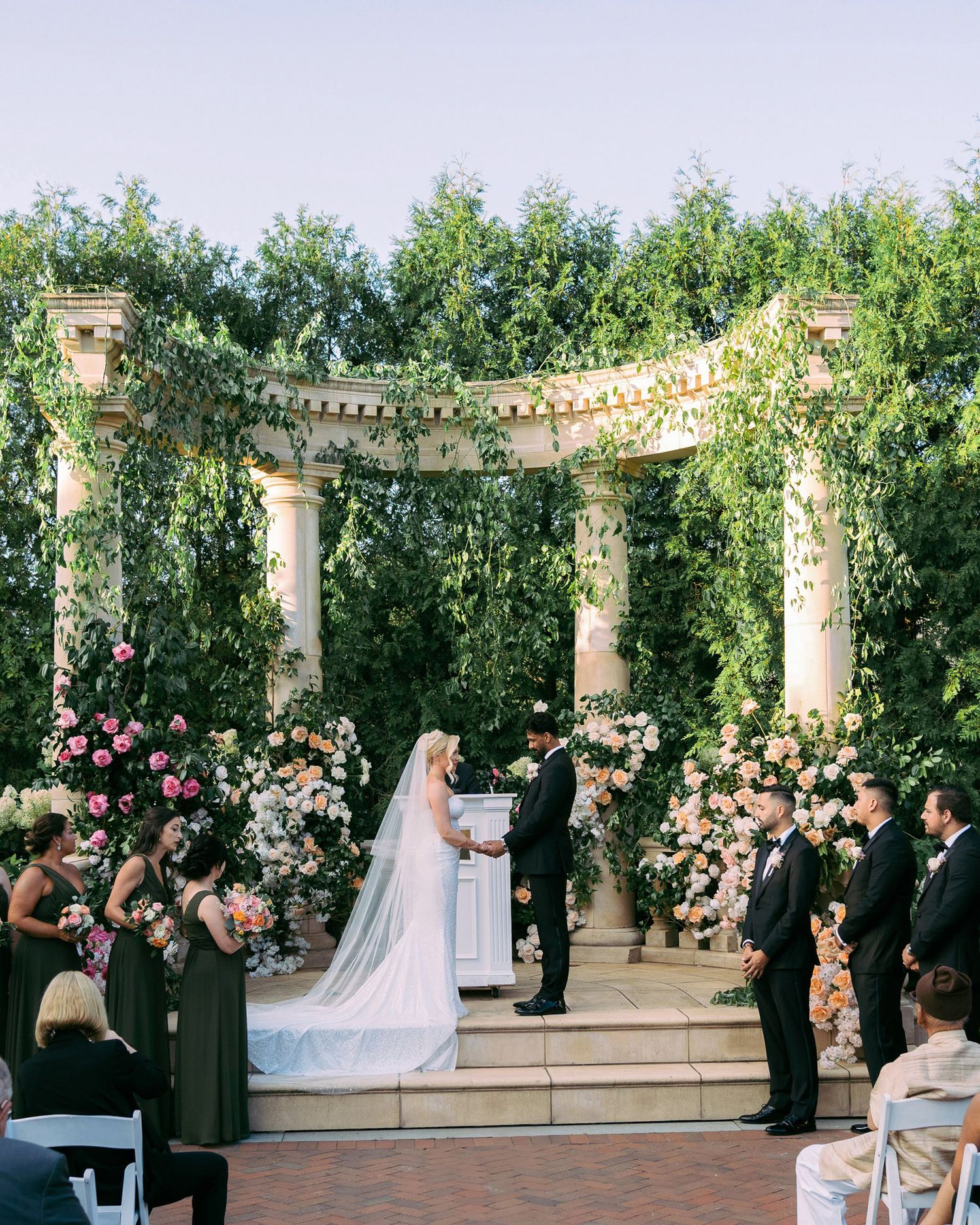 A panoramic view of an elegant outdoor wedding ceremony with floral arrangements and pillars in a garden setting.