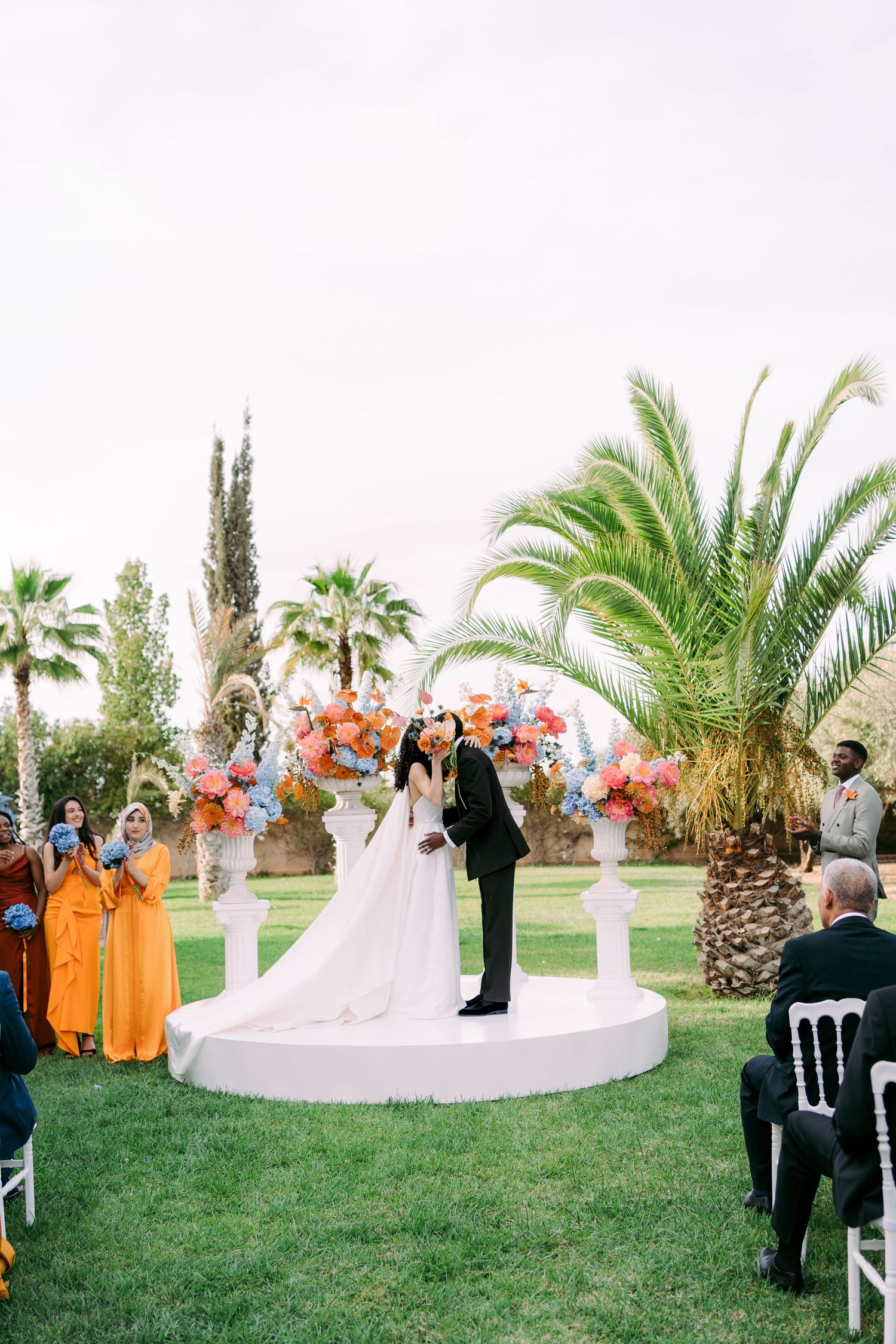 Outdoor wedding ceremony under palm trees with orange-clad wedding party and white floral arrangements.