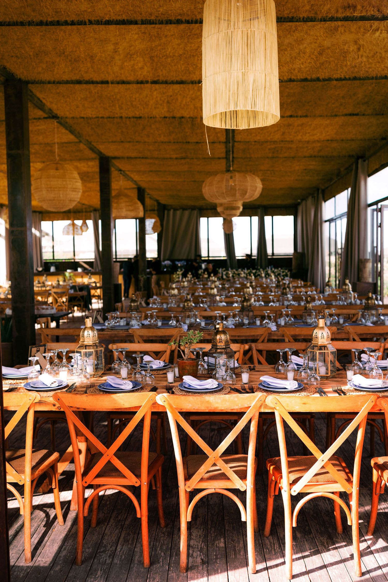 Long wooden dining tables set with place settings and orange cross-back chairs in a rustic reception venue.