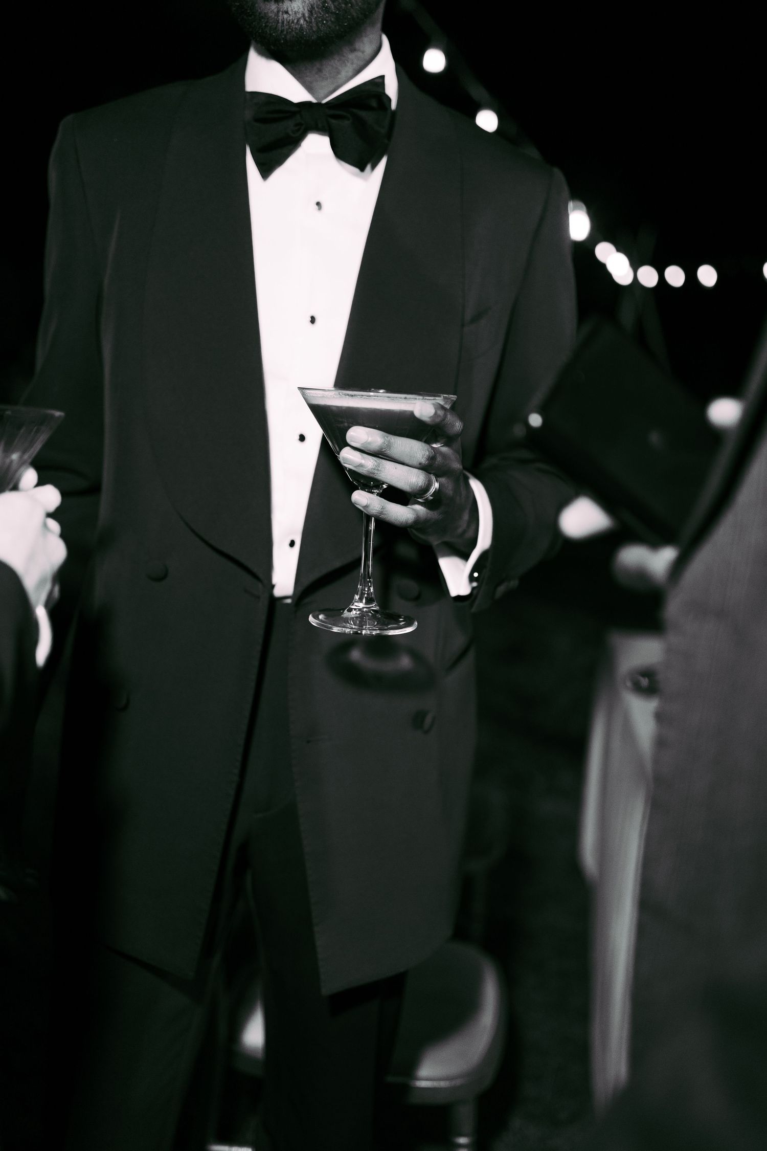 Men in tuxedos hold cocktail glasses at an elegant black tie event under dim lighting.