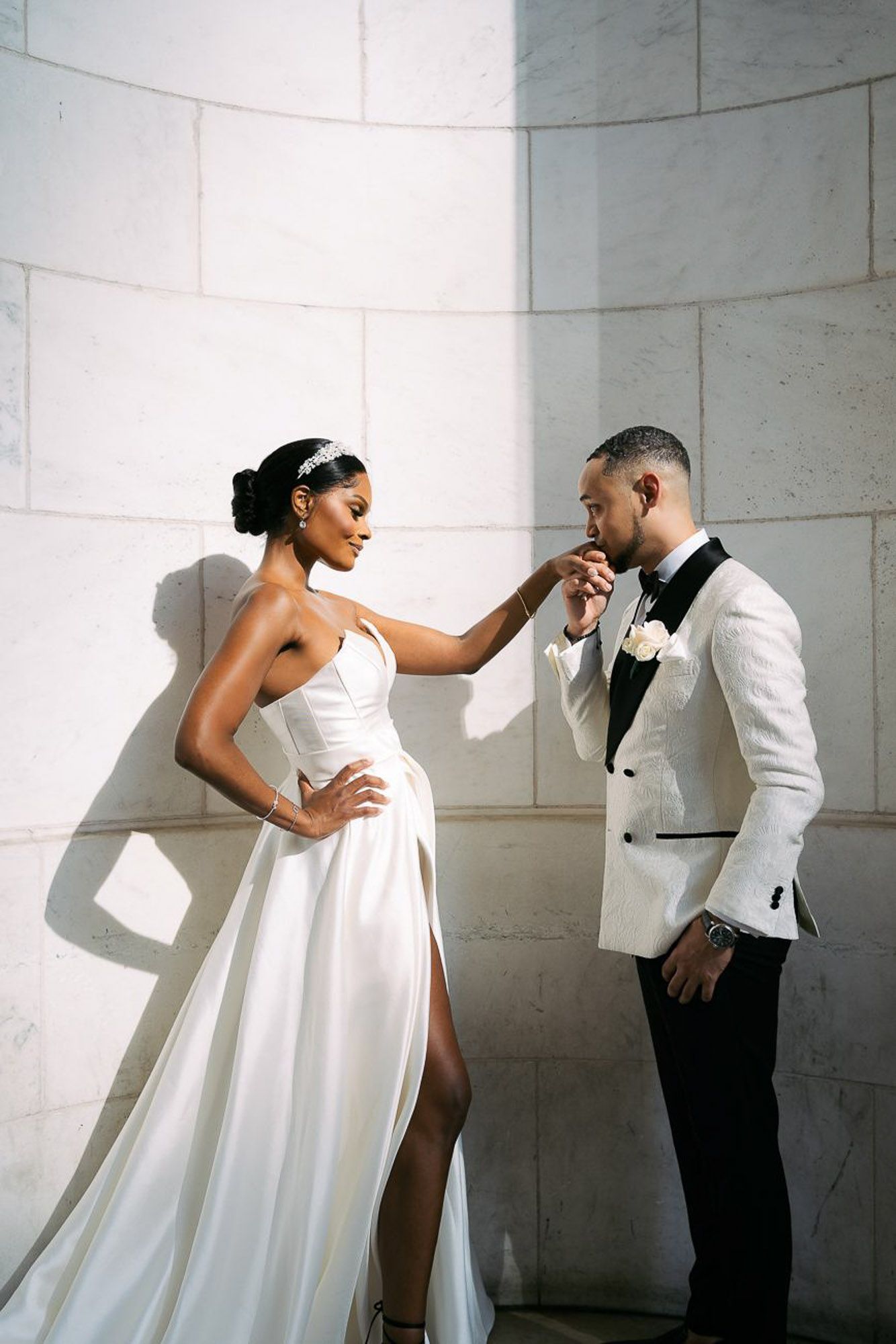 A romantic wedding moment between a couple in elegant white attire against a stone wall with dramatic lighting.
