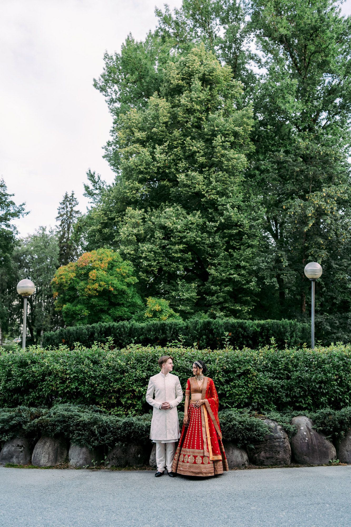 A couple in traditional Indian wedding attire poses together in a garden setting with decorative lamp posts.