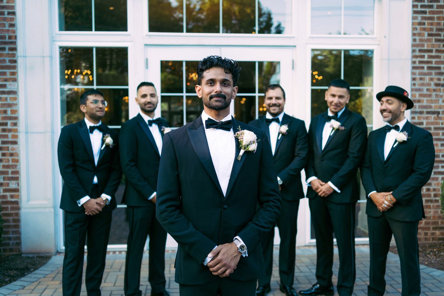 A wedding party in black tuxedos stands in a line against a brick building with white windows.