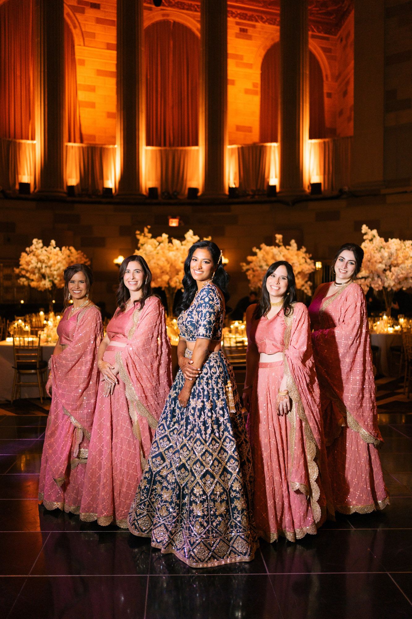 Traditional Indian wedding party wearing pink saris and a navy blue lehenga pose in an elegant venue with warm lighting.