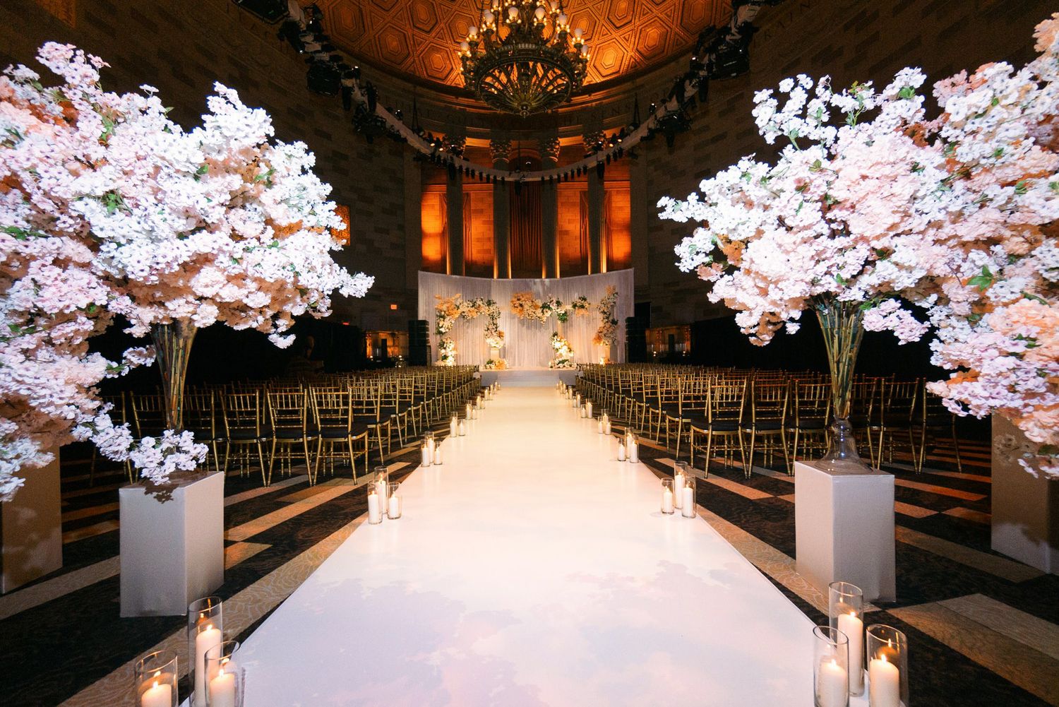 Indoor wedding ceremony venue with white aisle runner and cherry blossom trees flanking the path under warm lighting.