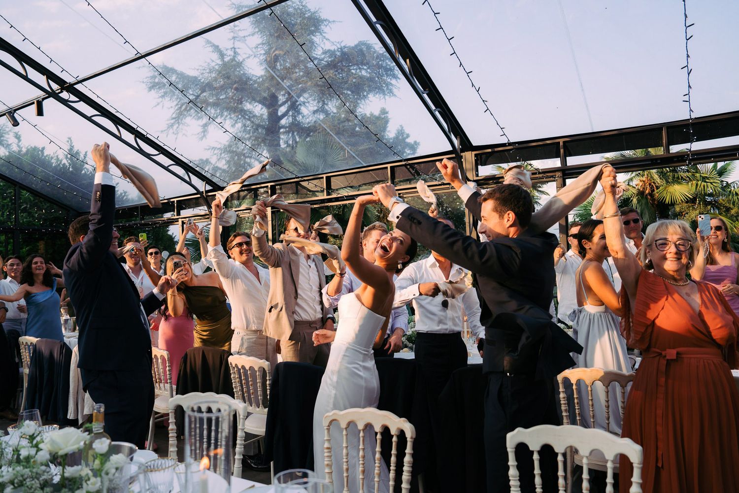 Wedding guests dance and celebrate under a clear tent during an outdoor evening reception.