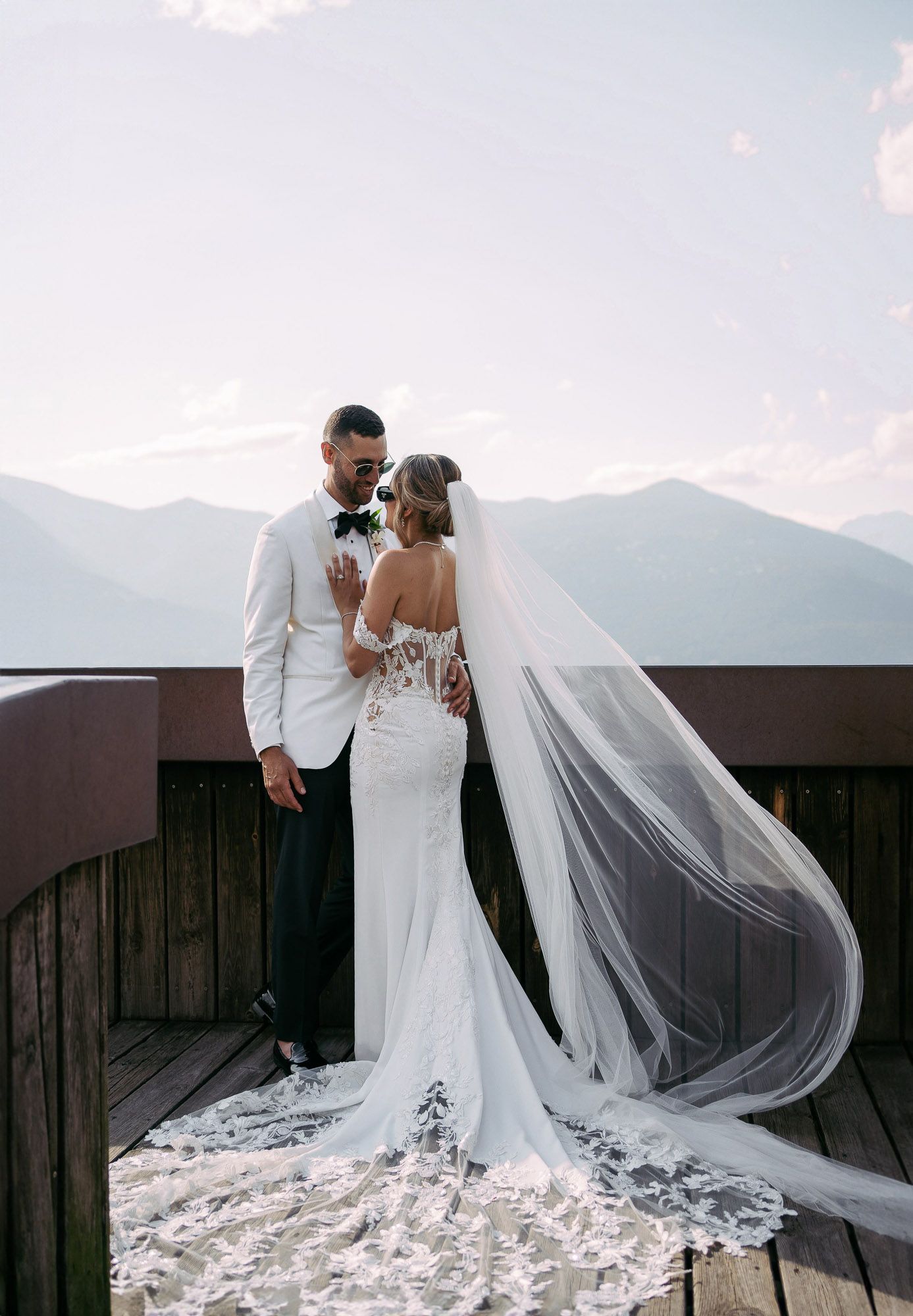 Wedding couple poses on mountain deck with dramatic veil flowing in breeze against scenic mountain backdrop.