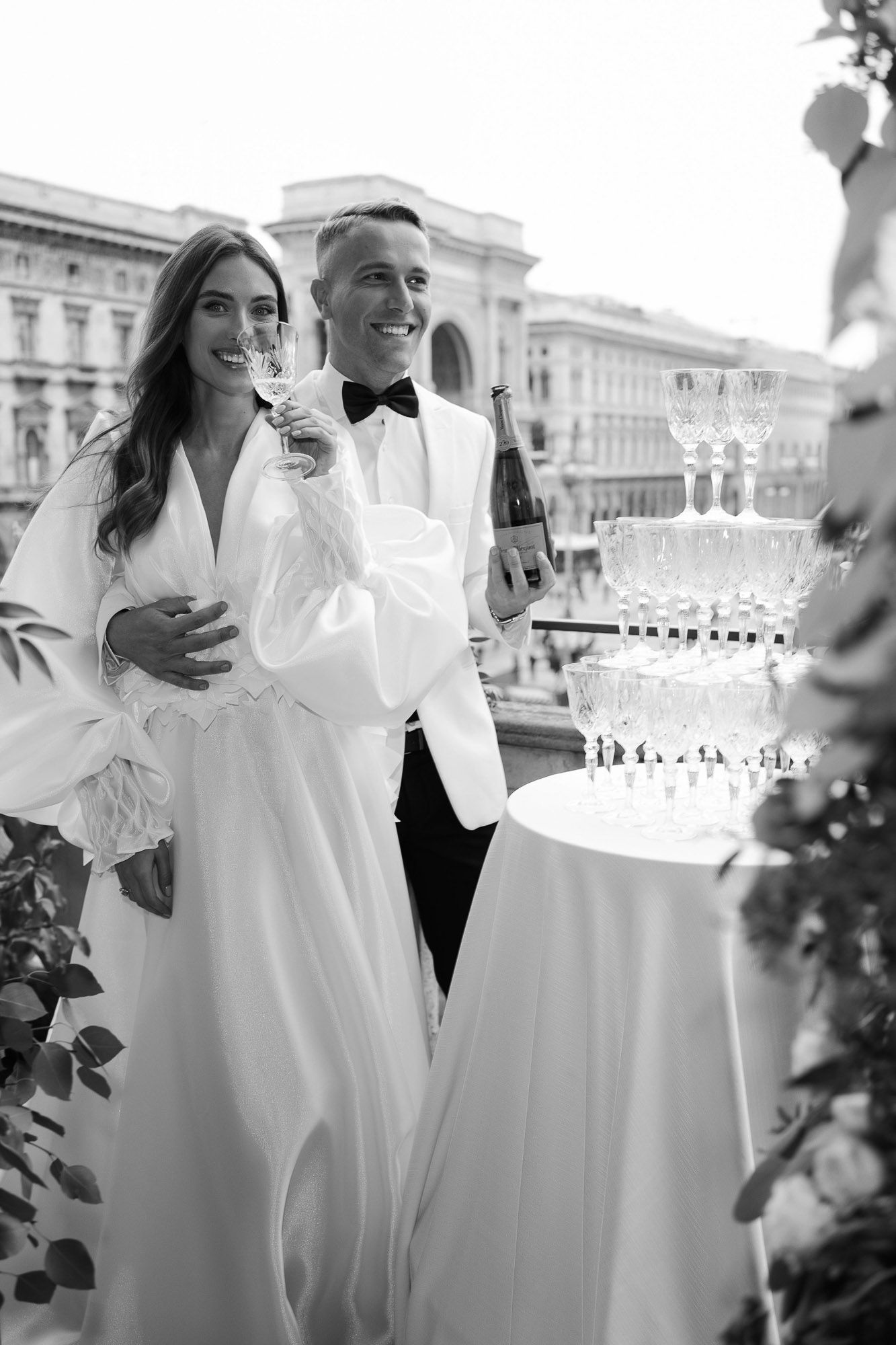 Guests in elegant white attire raise champagne glasses during an outdoor celebration at a formal venue.