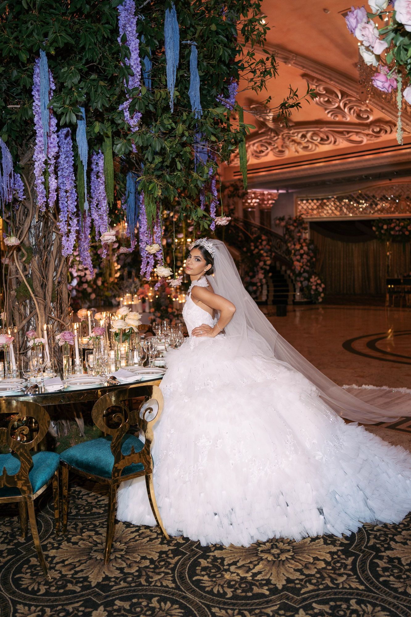 A flowing white wedding gown with a dramatic train is photographed in an ornate ballroom with purple floral decor.