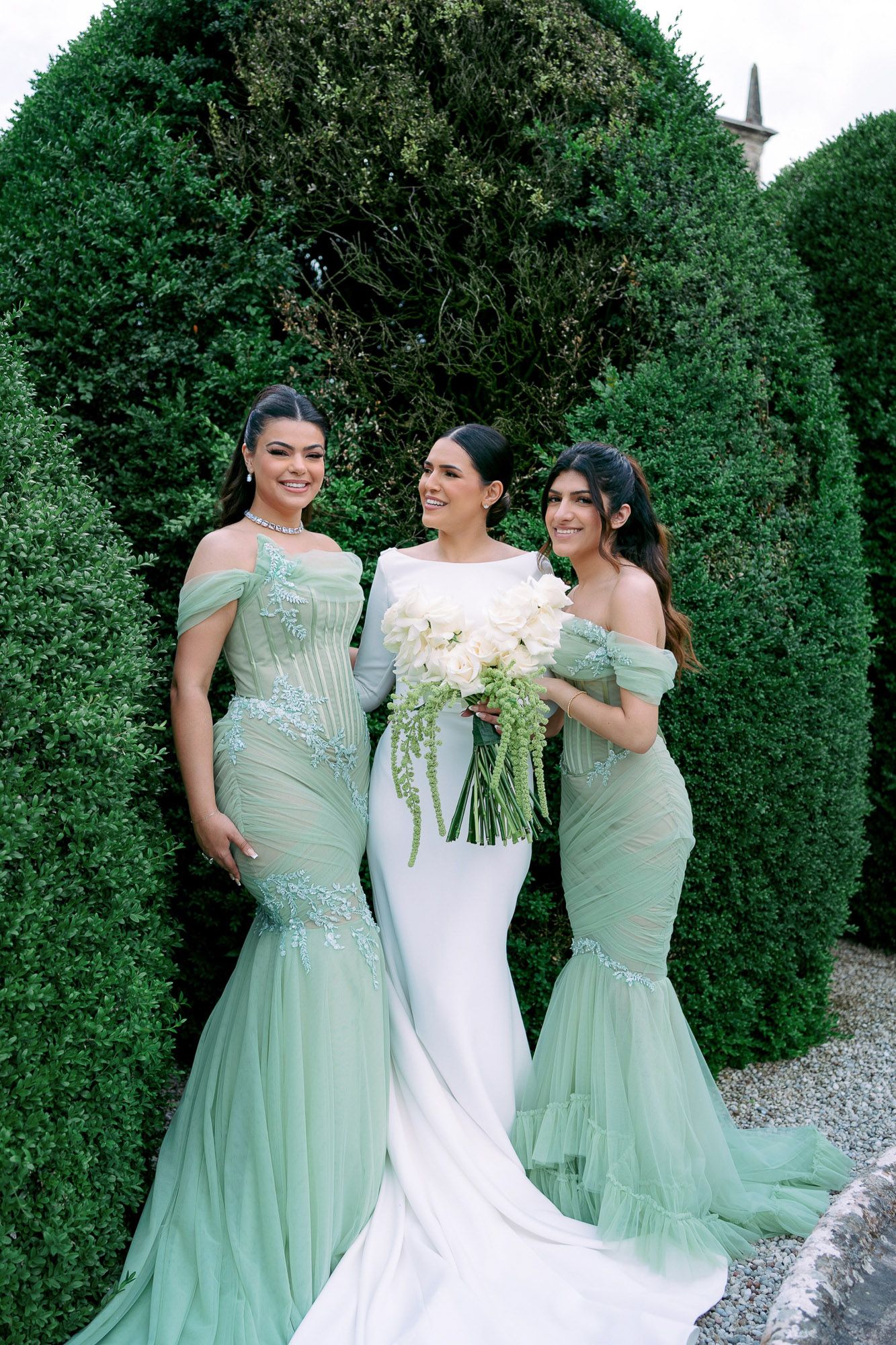 A bride in white gown stands between two bridesmaids in mint green dresses against a lush hedge backdrop.