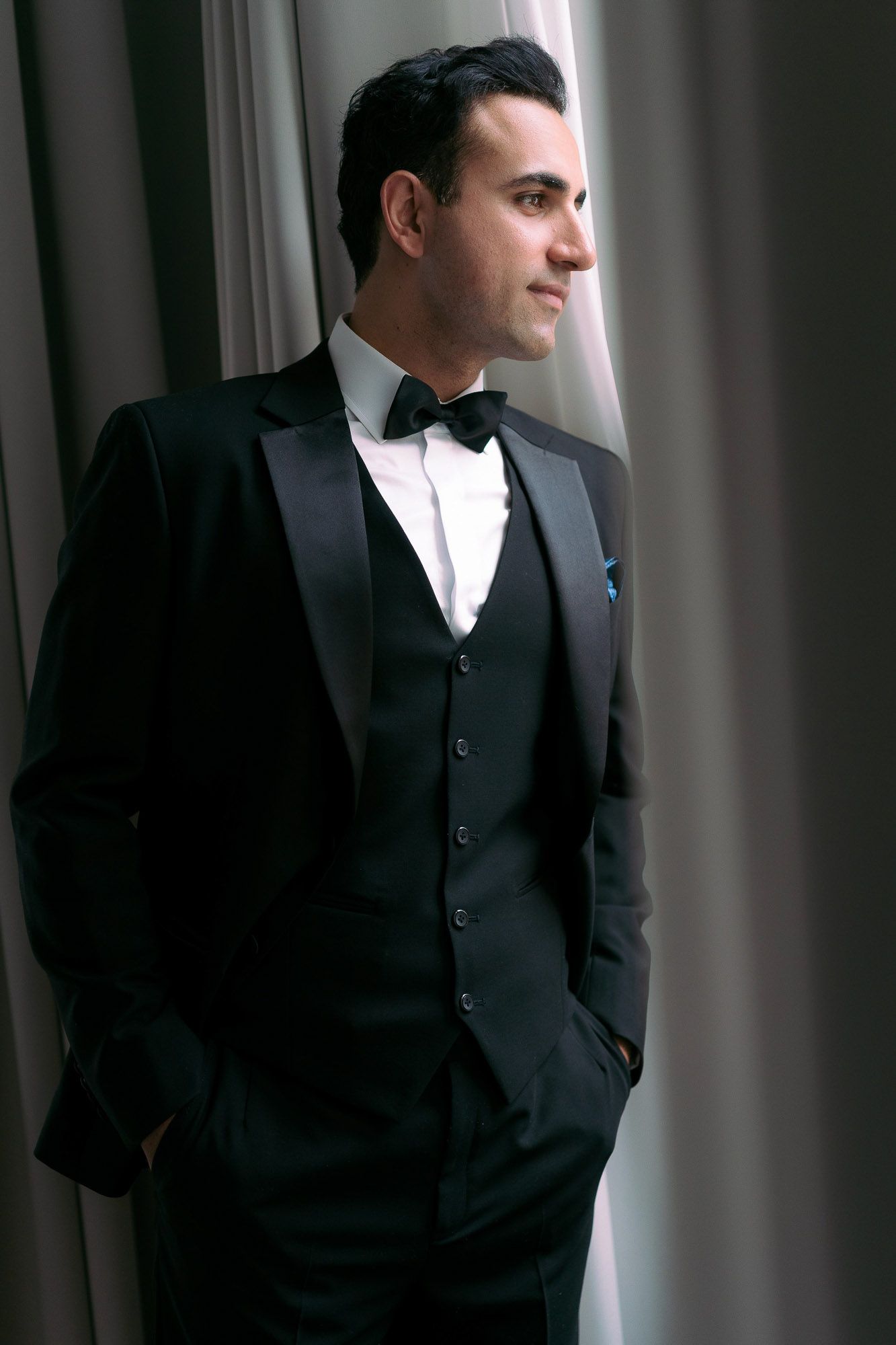 A man in a formal black suit and white shirt stands looking contemplatively out a window in dramatic lighting.