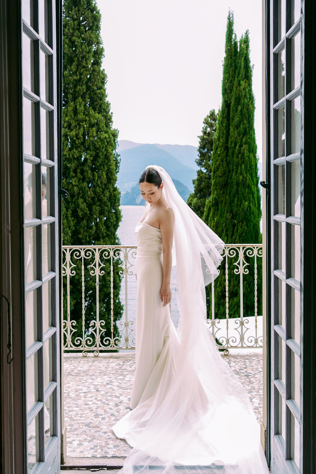 Elegant bride in flowing white veil stands by French doors overlooking cypress trees and mountains in romantic setting.