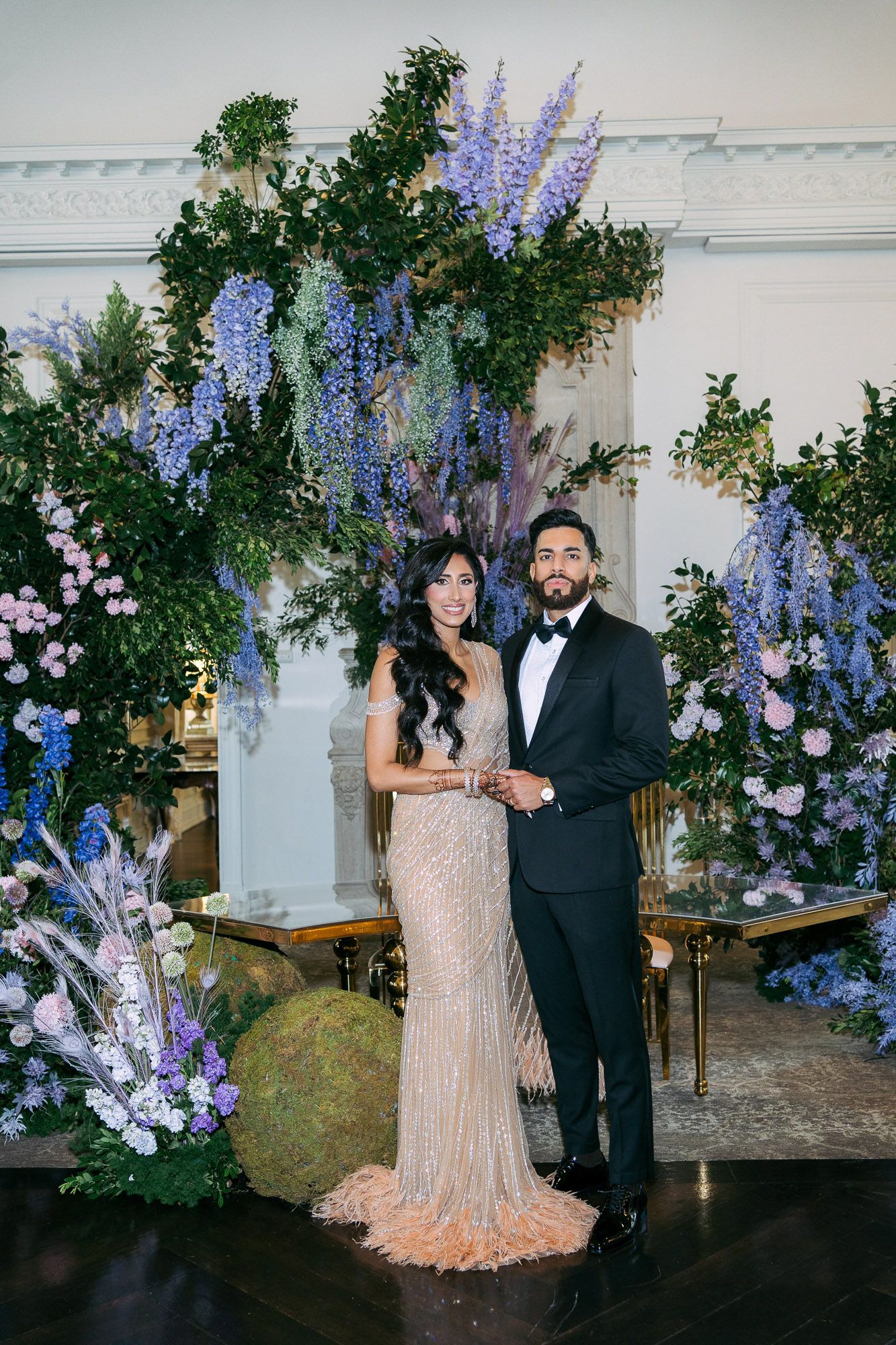 Elegant couple poses in front of romantic floral backdrop with purple wisteria and greenery at evening reception.