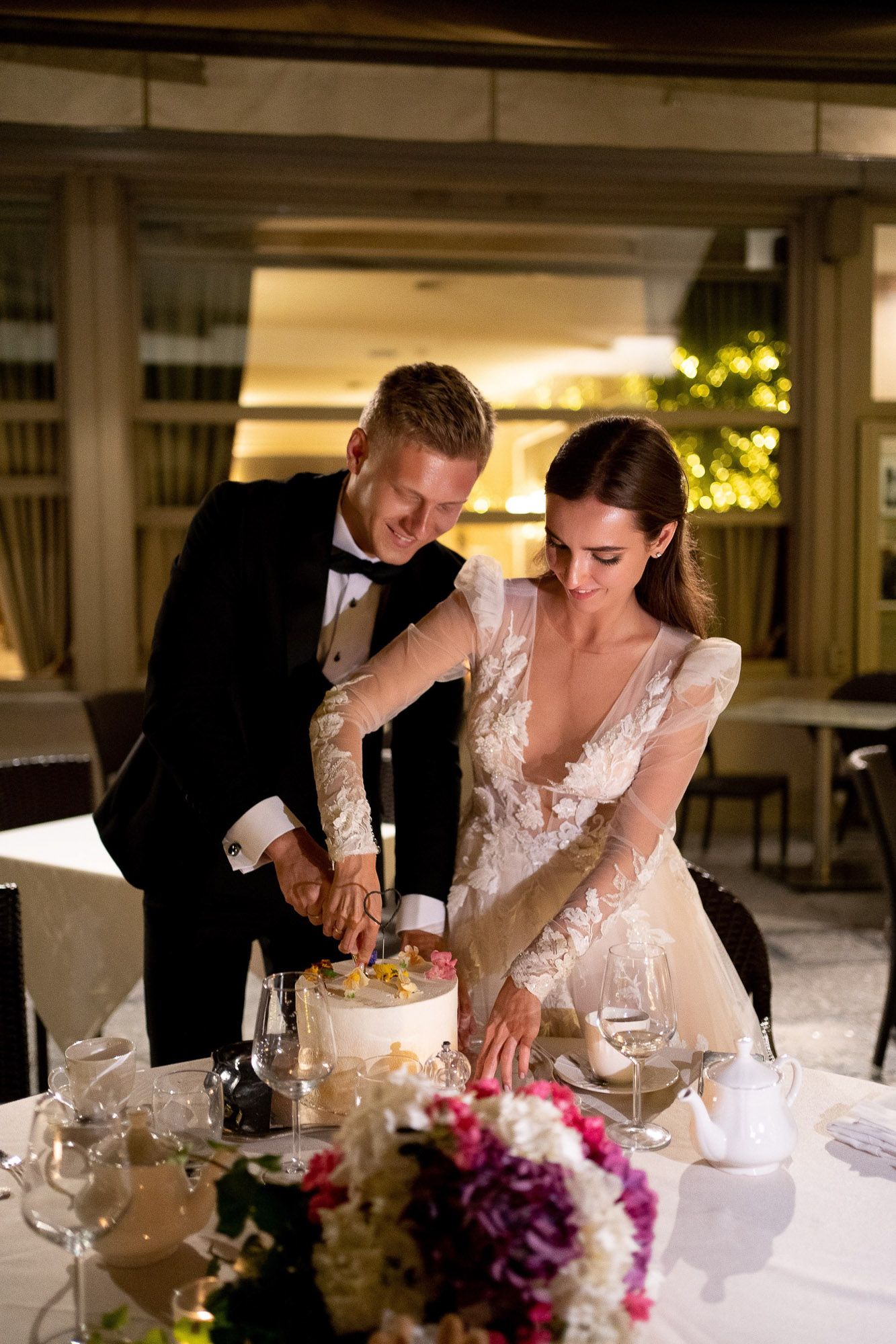 A couple in wedding attire shares an intimate moment while cutting their wedding cake during a reception dinner.