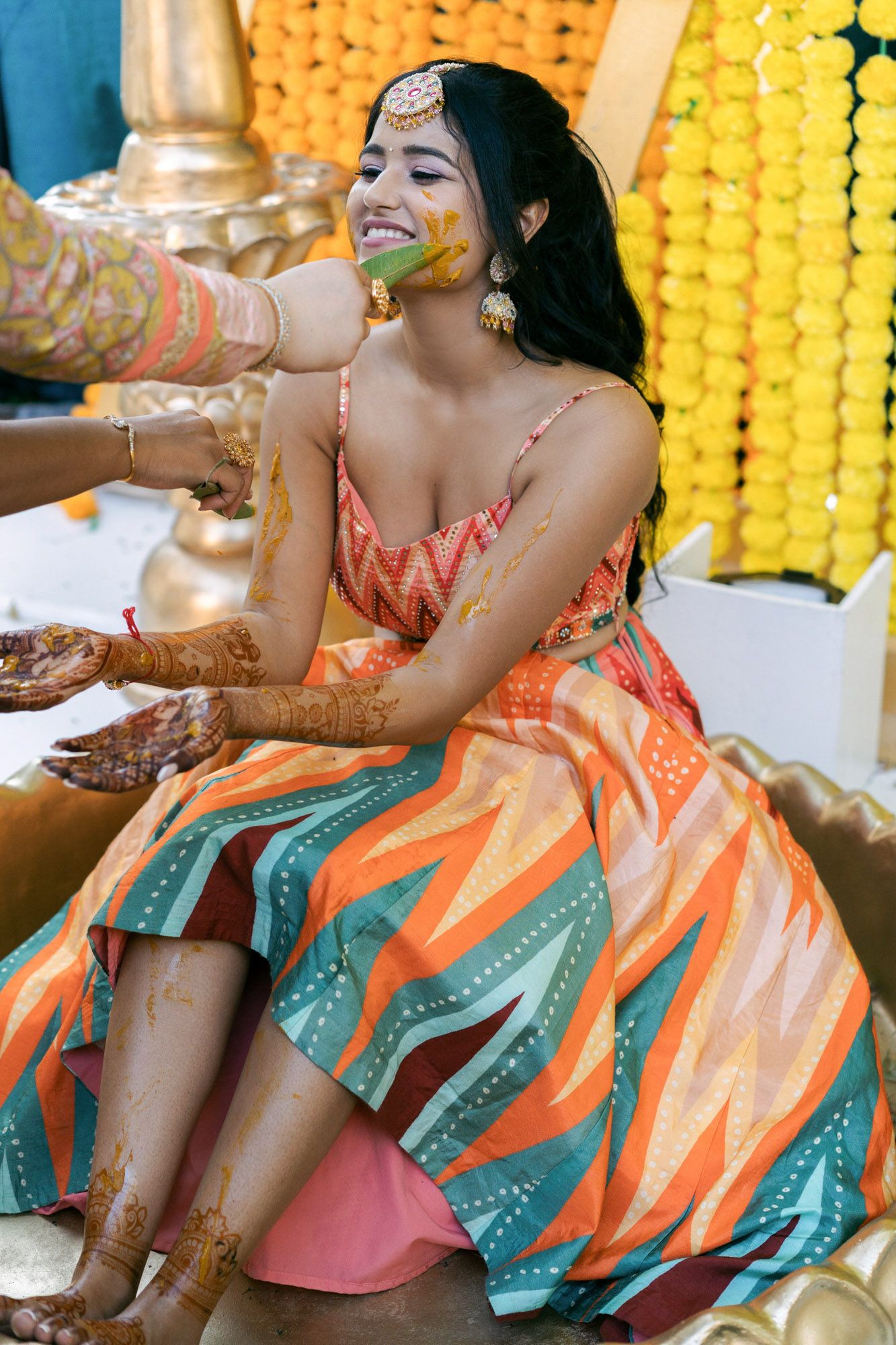 A person in a colorful orange and teal dress participates in a traditional haldi ceremony with yellow decorations.