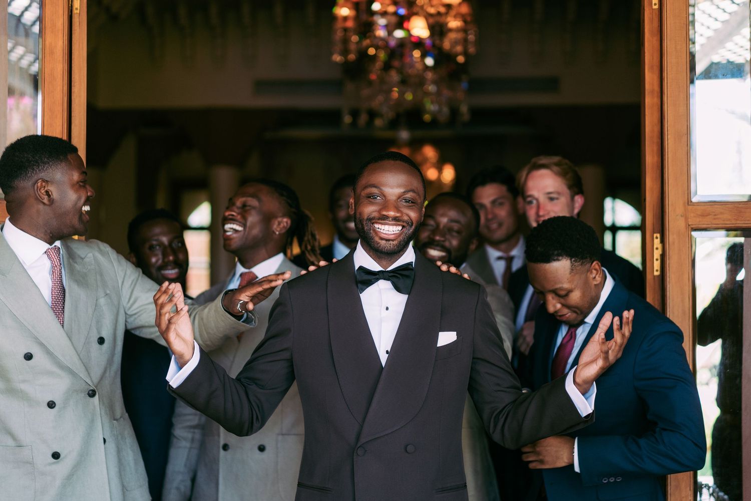 Groomsmen in formal suits and tuxedos gather for a celebratory wedding photo indoors.