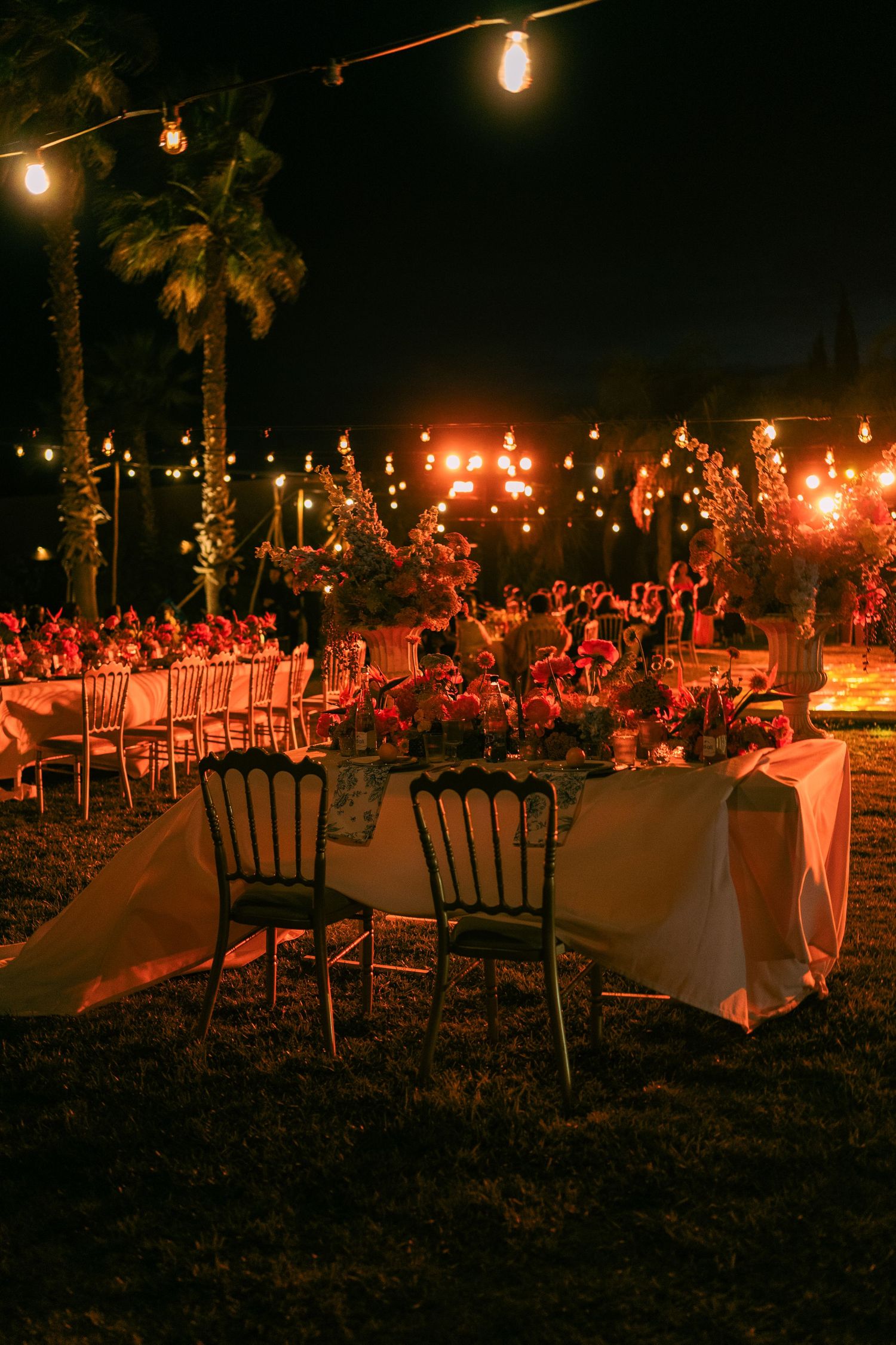 Outdoor wedding reception setup with string lights and elegant table settings under palm trees.