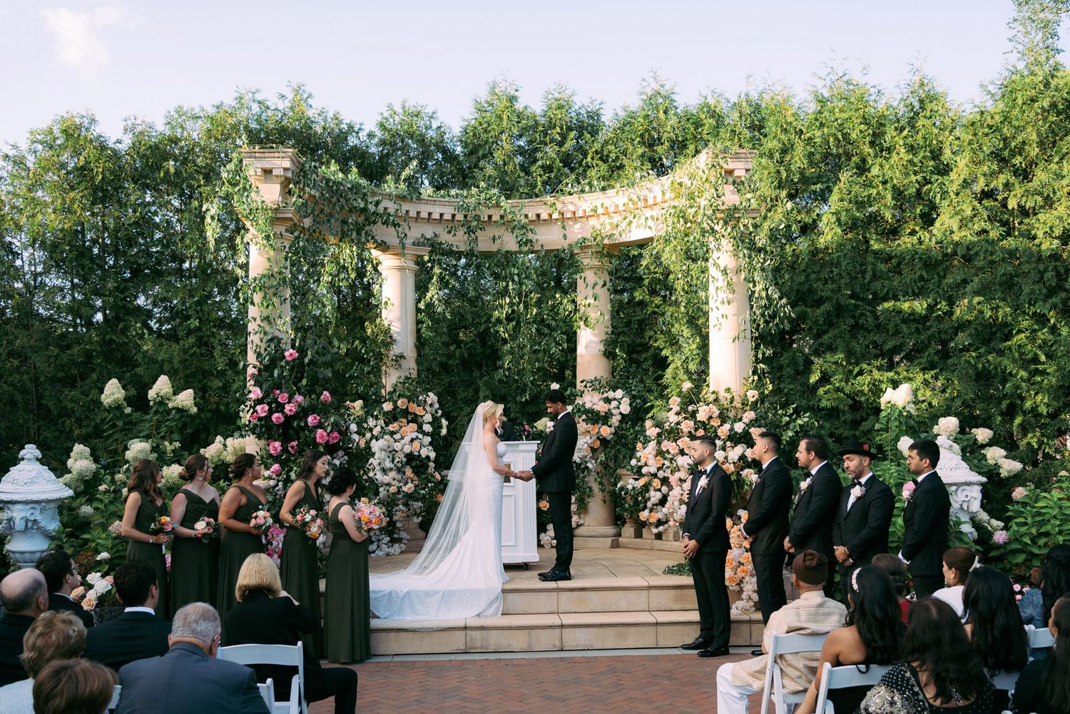 An outdoor garden wedding ceremony takes place under a beautiful floral-adorned pergola with guests seated on white chairs.