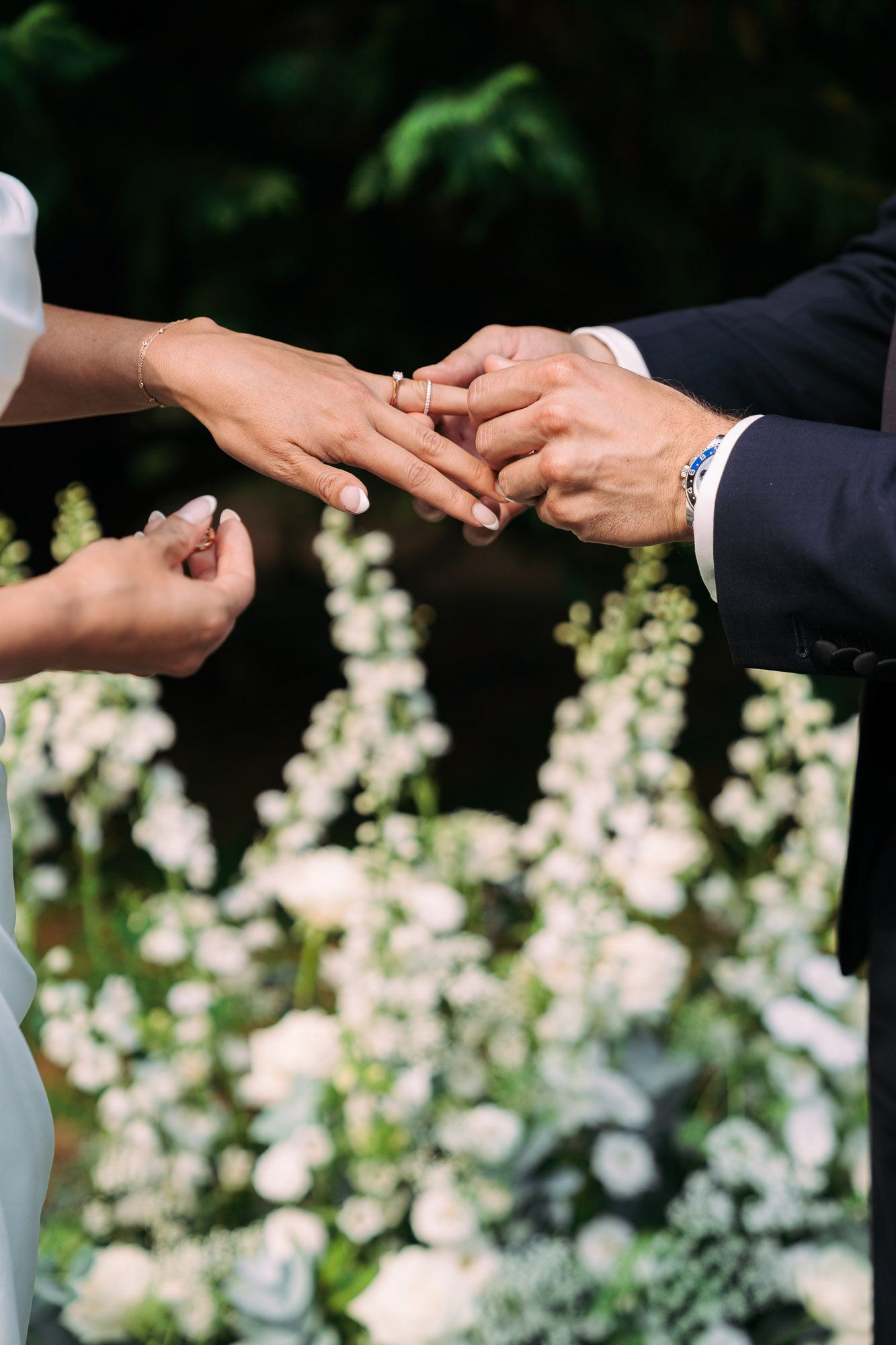 Close-up shot of hands exchanging wedding rings during outdoor garden ceremony with white flowers in background.