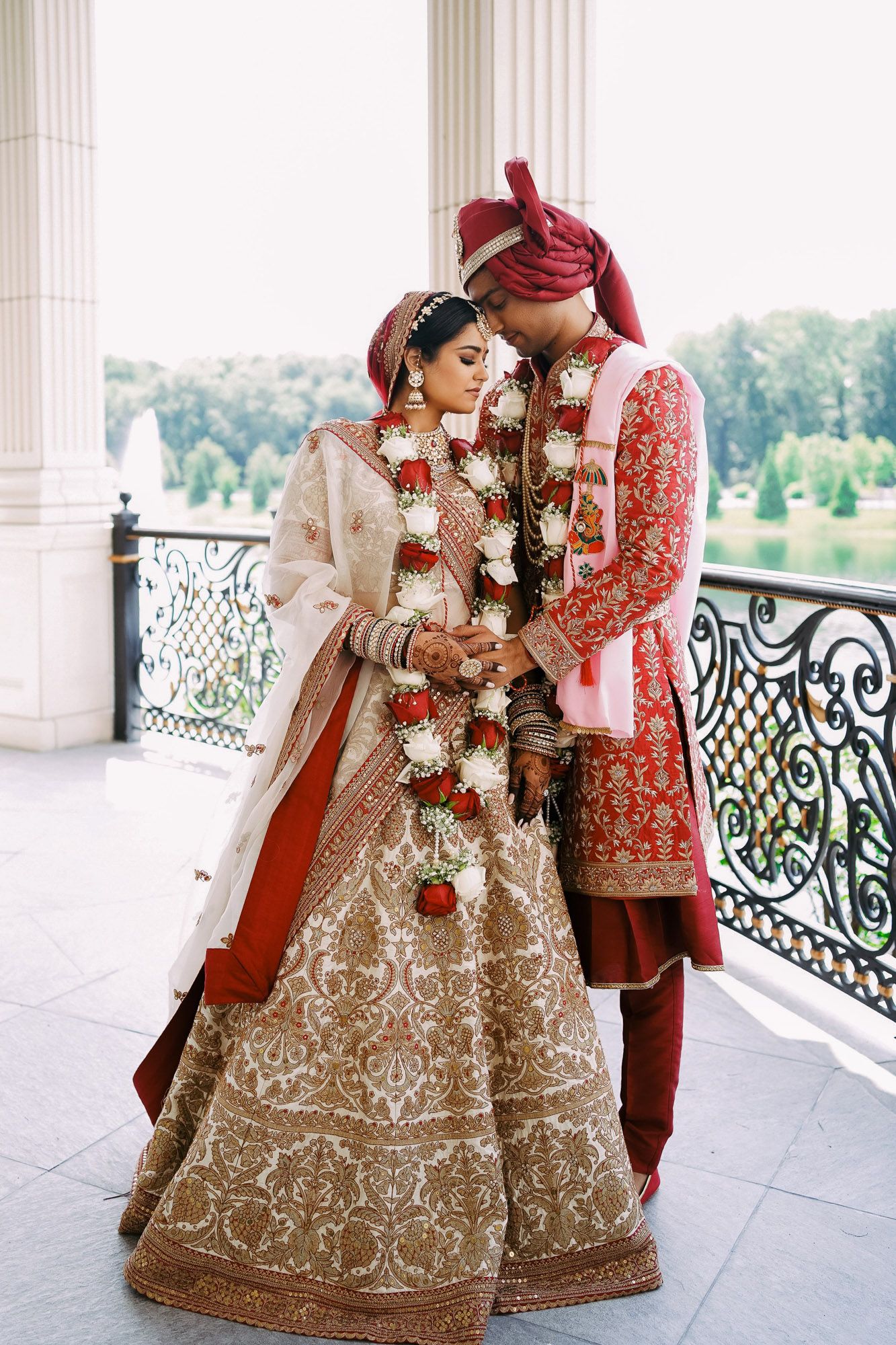 Traditional Indian wedding couple in elegant red and gold attire pose together on an ornate balcony with scenic views.
