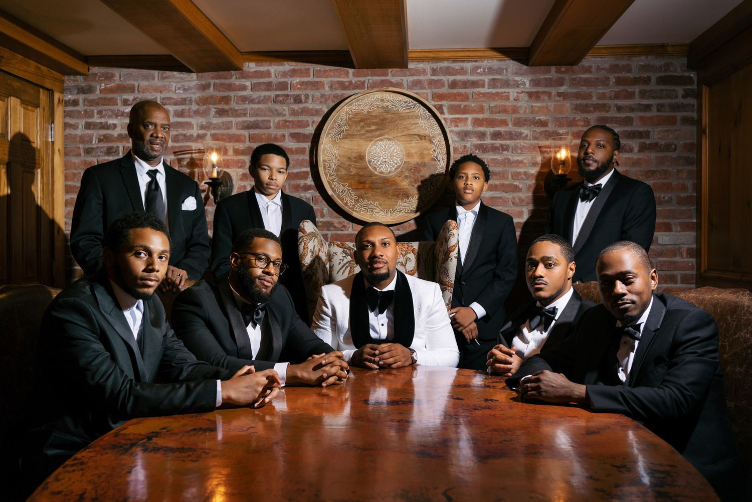 A group in formal attire sits at a long wooden table in a rustic brick-walled room during a celebratory gathering.
