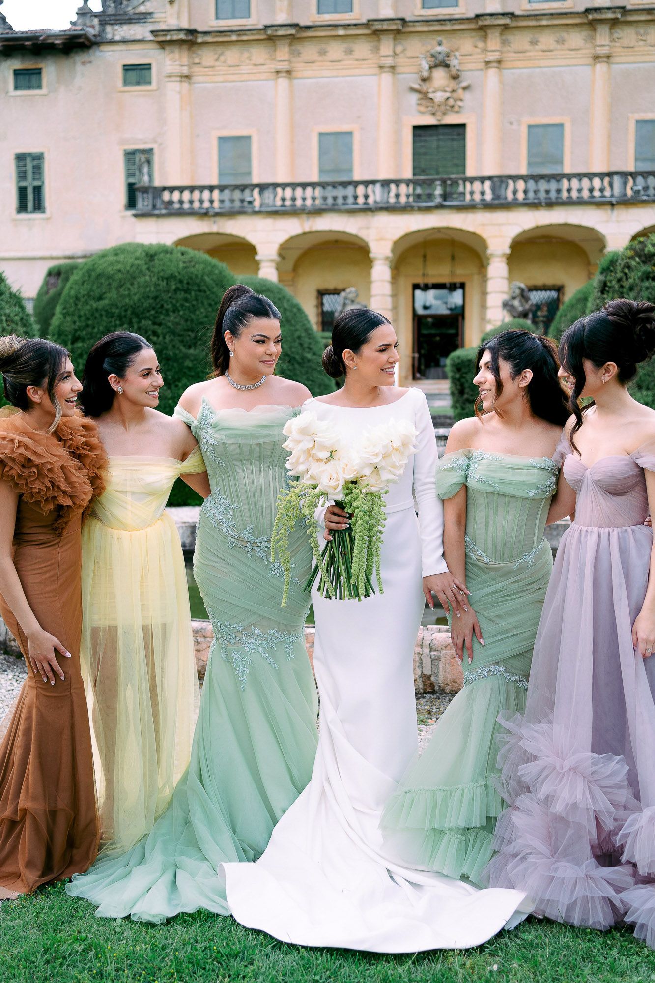 A row of bridesmaids in pastel mint green, lavender, and brown dresses pose together at an elegant venue.