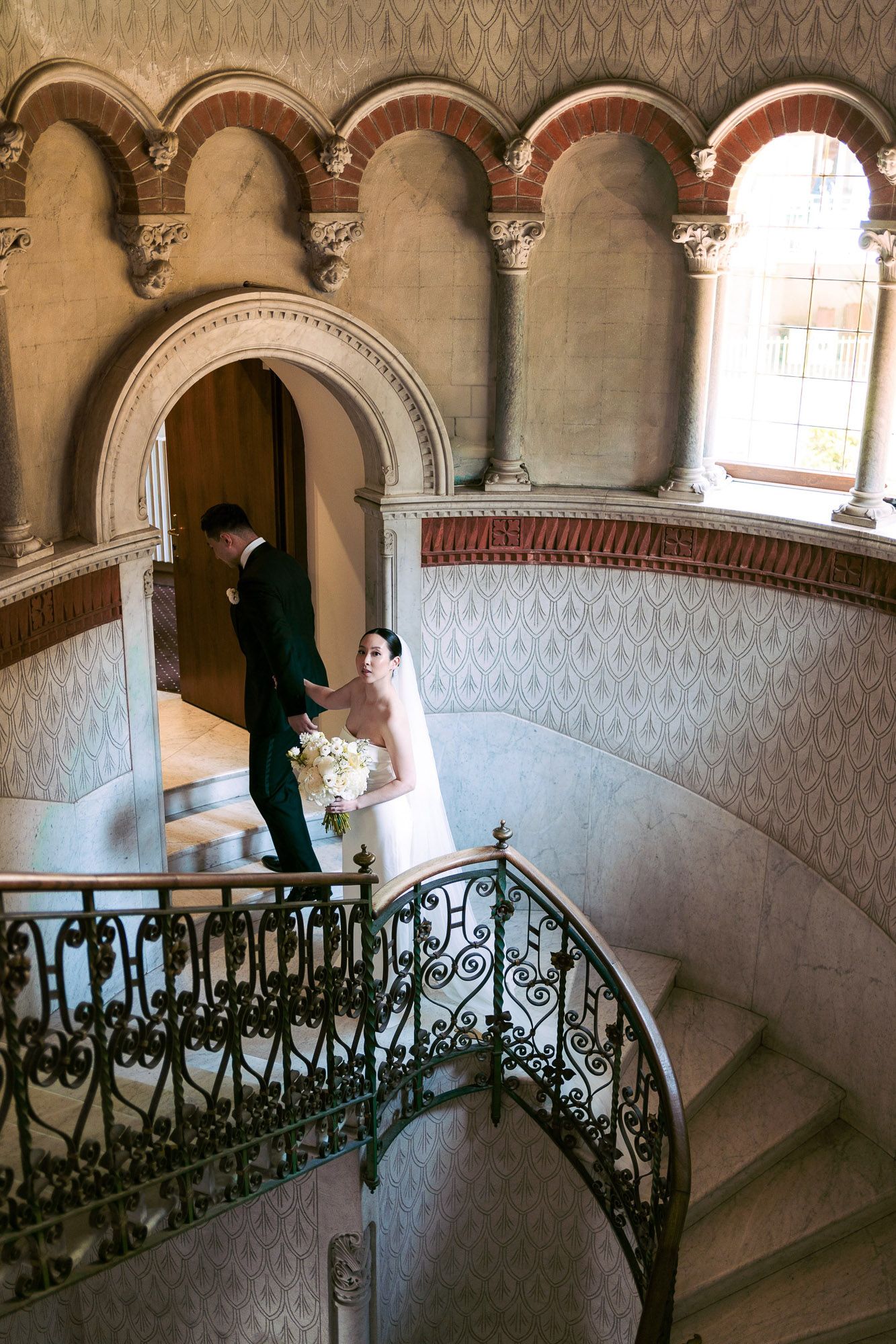 Elegant curved staircase with ornate iron railings and arched columns in a historic mansion interior.