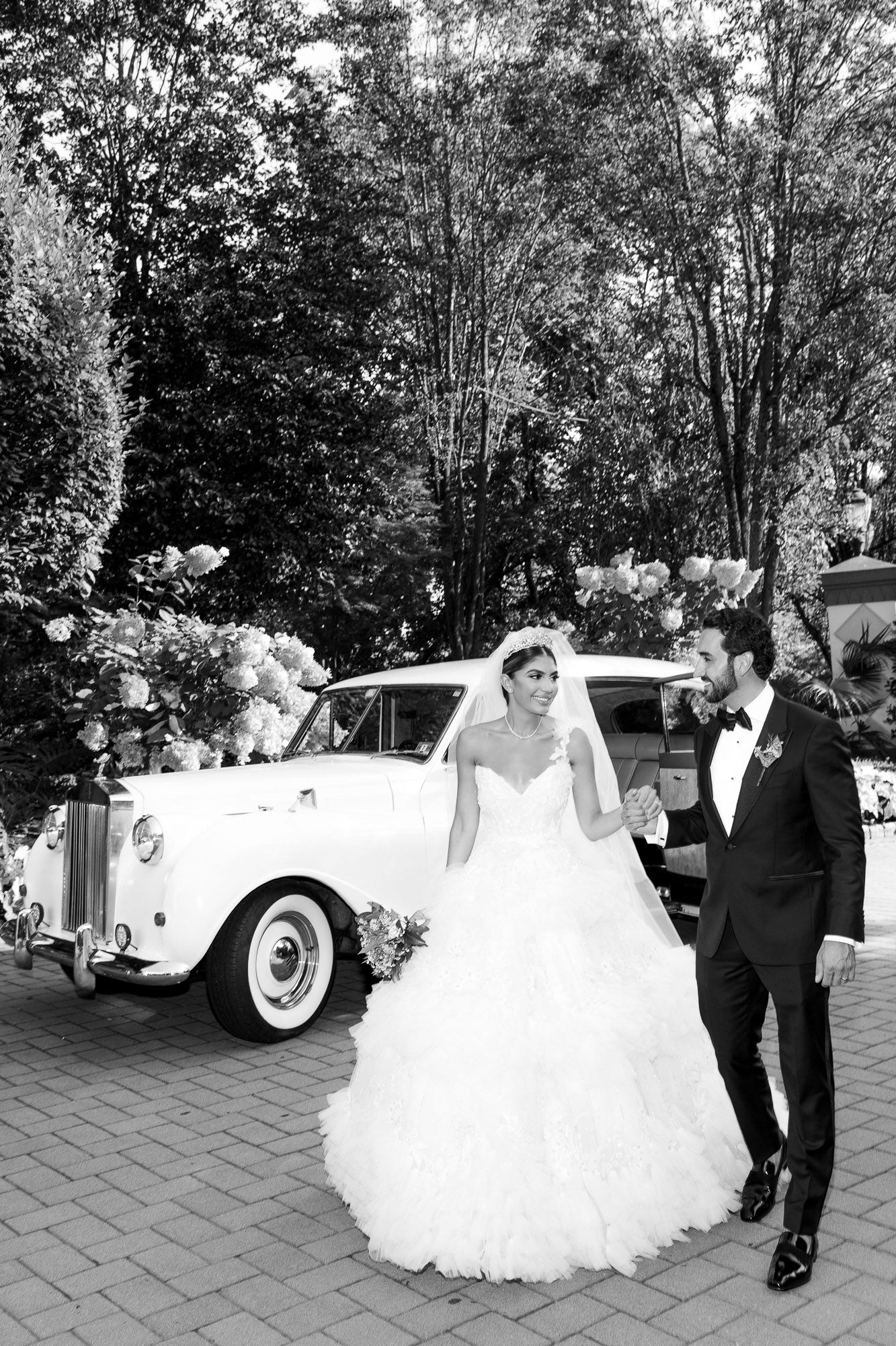 A wedding party poses near classic vintage white cars on a stone paved courtyard during an elegant outdoor celebration.